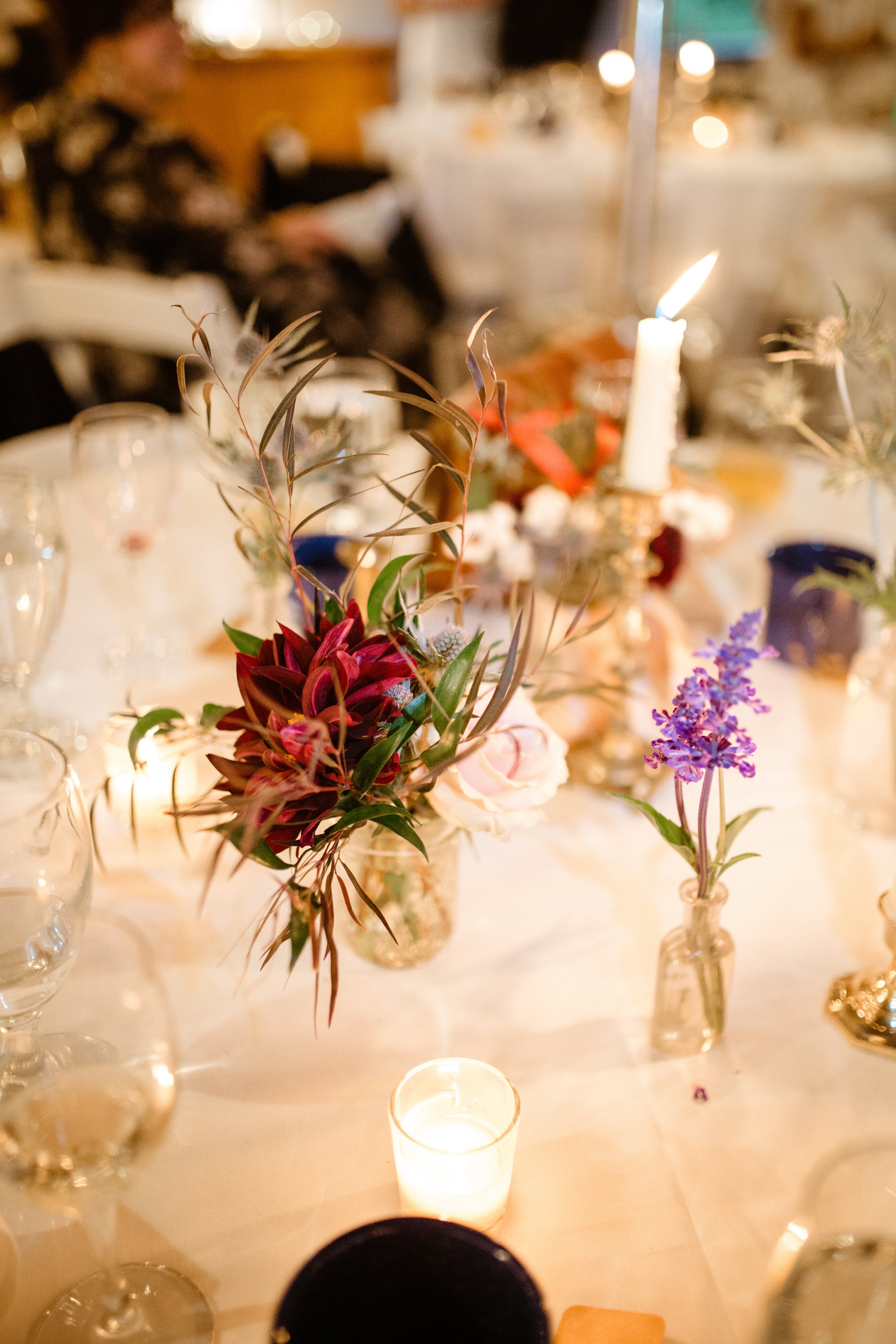 A table decorated with flowers and candles, with a blurred background of people and warm lights.