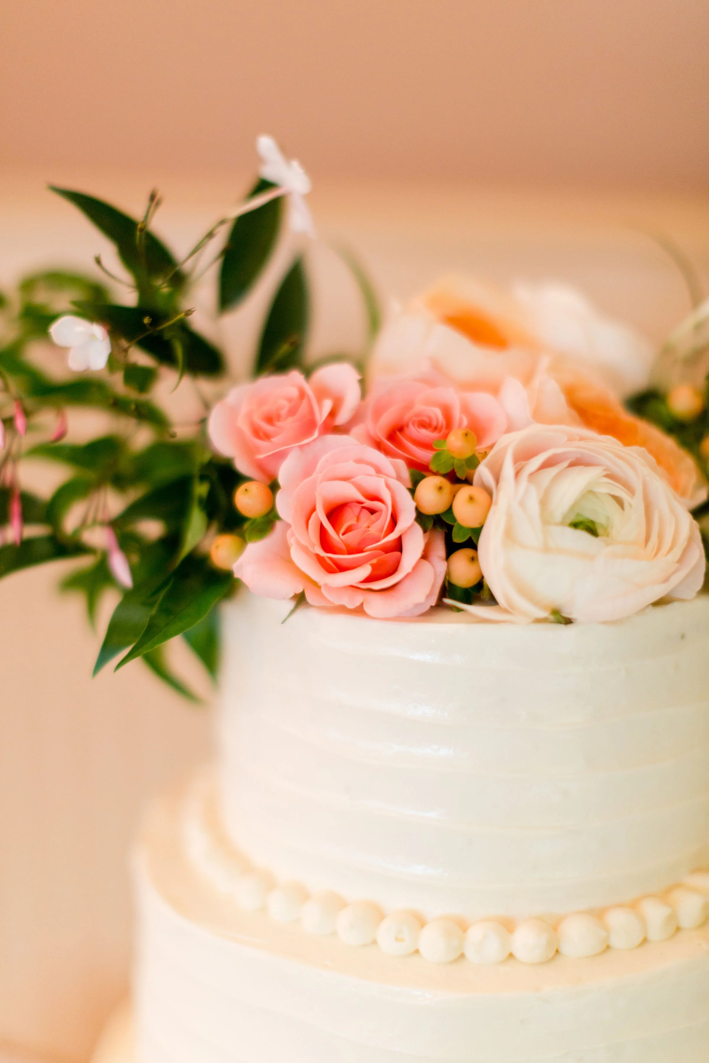 A white cake decorated with pink and white roses, yellow berries, green leaves, and a white flower on top, with a row of white fondant balls around the base.