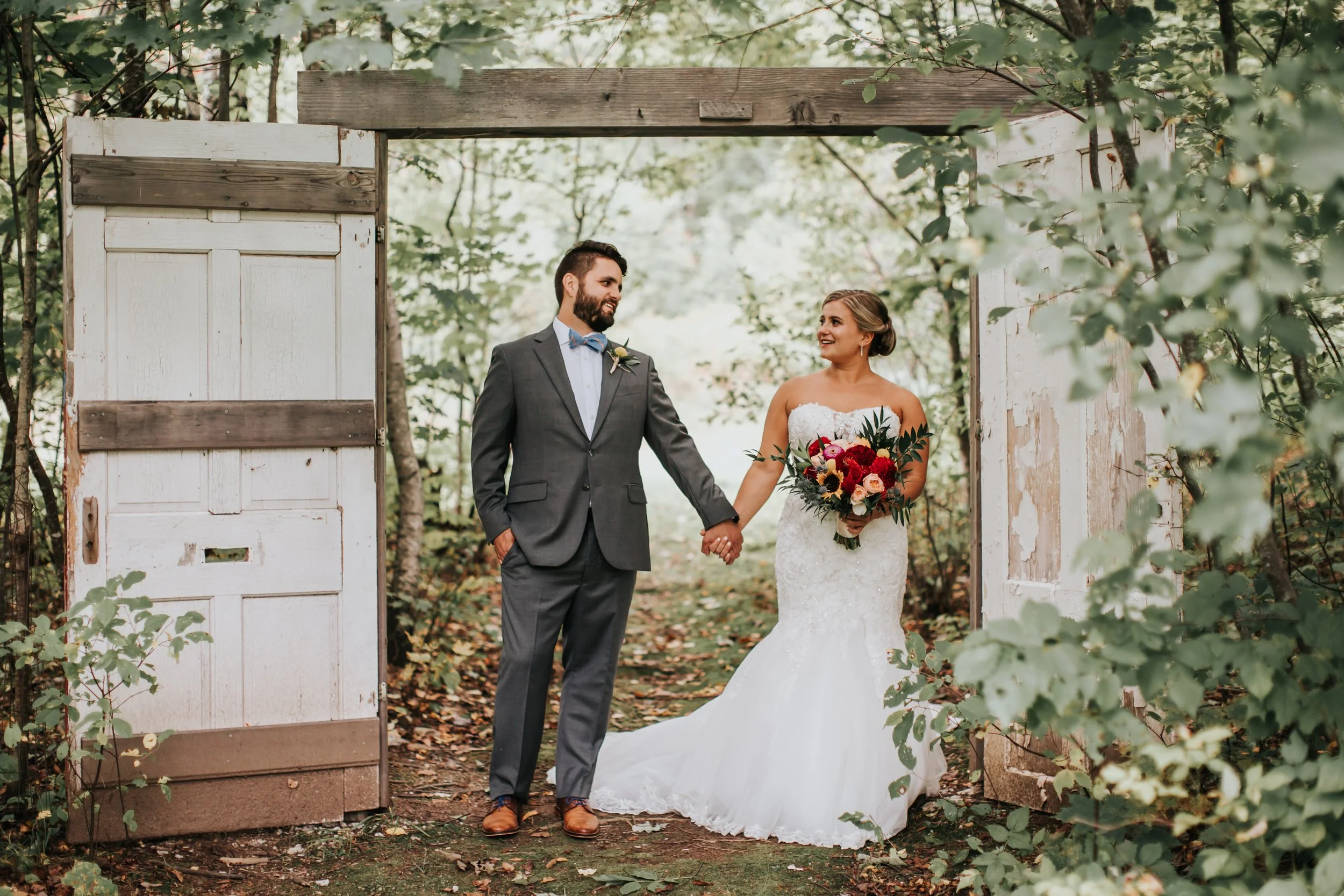 A groom and bride standing outside in a wooded area during their wedding, holding hands and smiling at each other. The groom is wearing a gray suit with a bow tie, and the bride is dressed in a strapless white wedding gown holding a bouquet of red, p