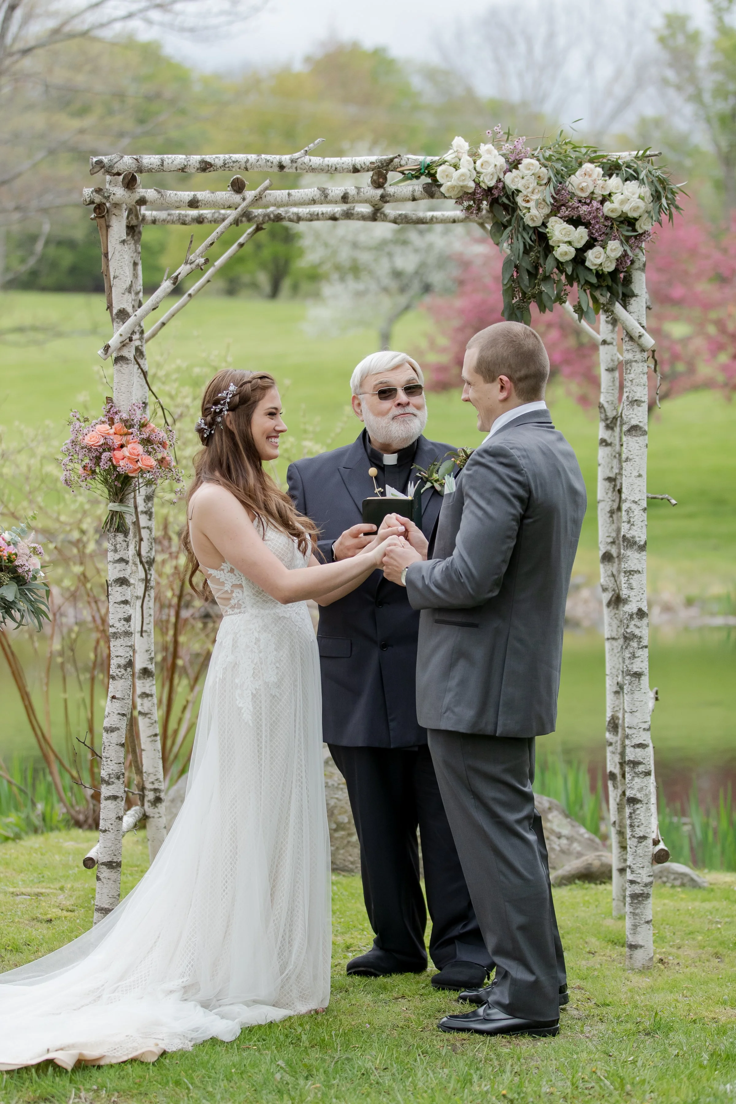 A couple getting married outdoors with a woman in a white lace wedding dress and groom in gray suit, holding hands and facing each other under a rustic wooden arch decorated with white and pink flowers, with an officiant in black robes standing betwe