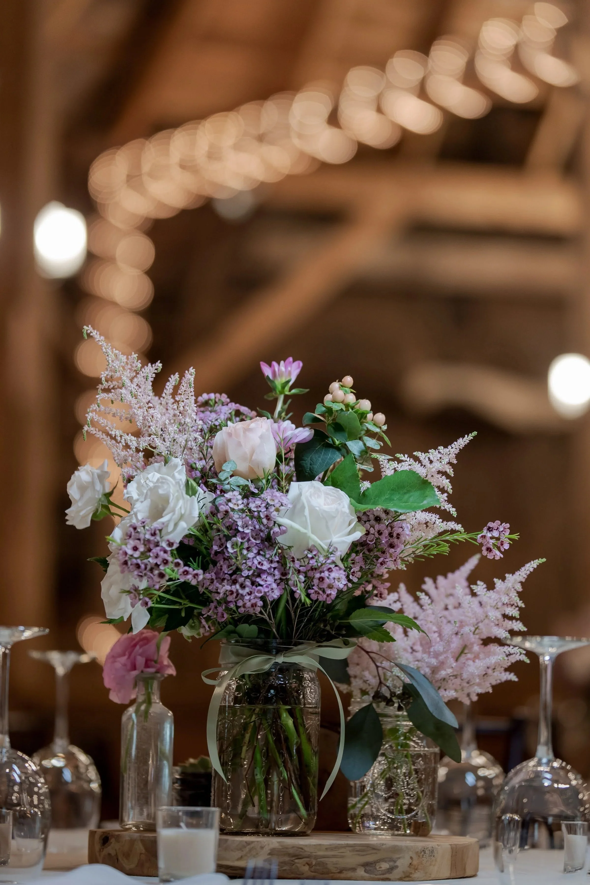 A floral centerpiece with pink and white flowers in a glass vase on a wooden slab, surrounded by glass votive candles on a table. The background features warm, blurred lights.