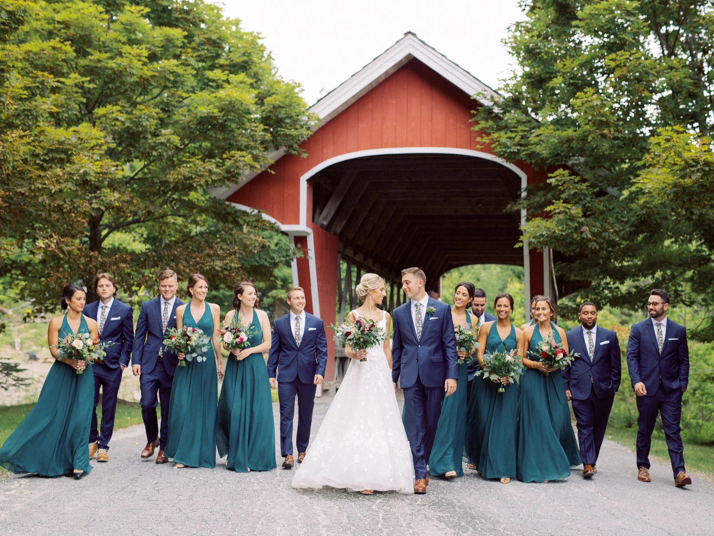 A wedding party walking outdoors on a path, with a red covered bridge and green trees in the background.