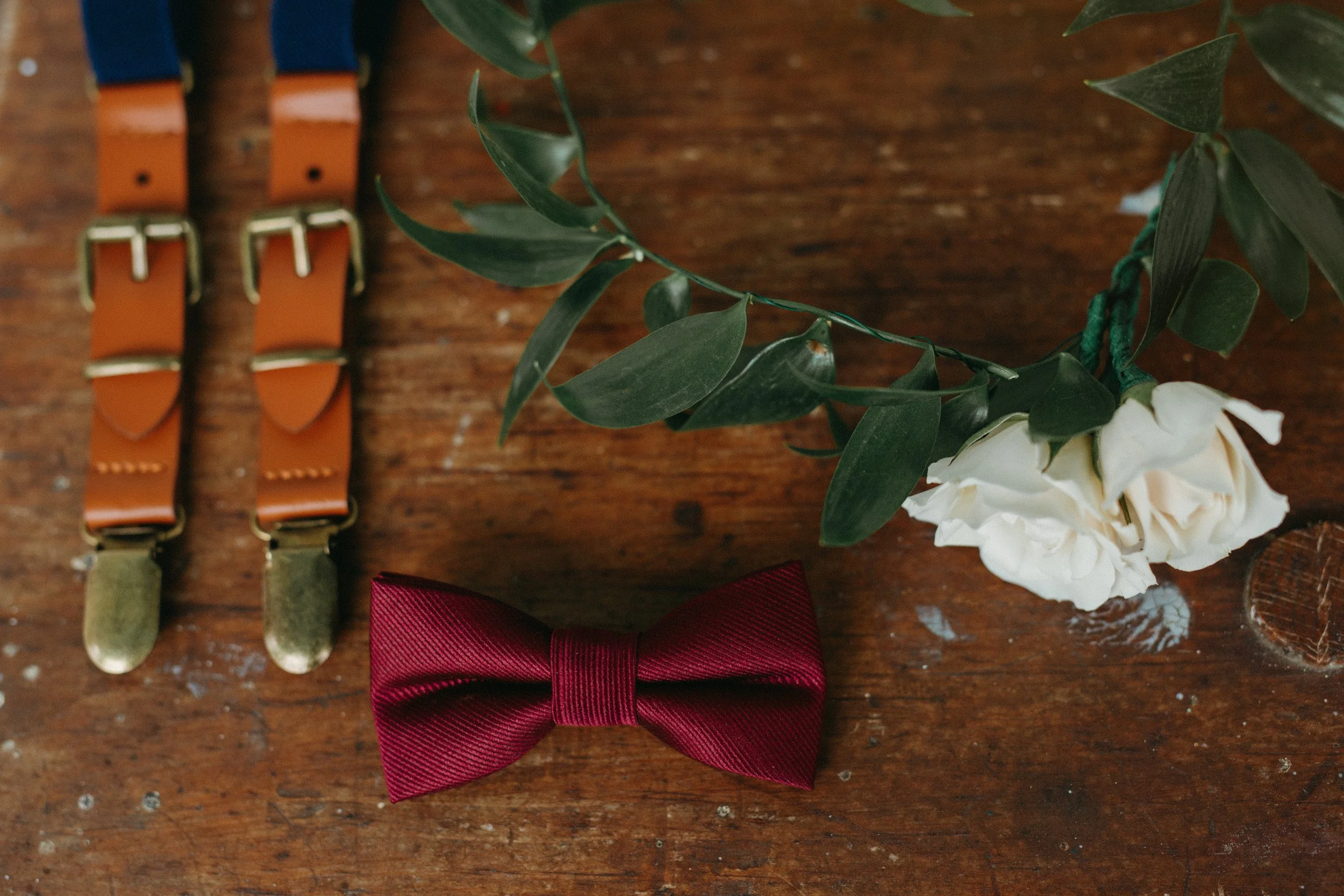 Close-up of a wooden surface with a pair of vintage-style suspenders, a white flower, a burgundy ribbon tied in a bow, and a leafy plant.