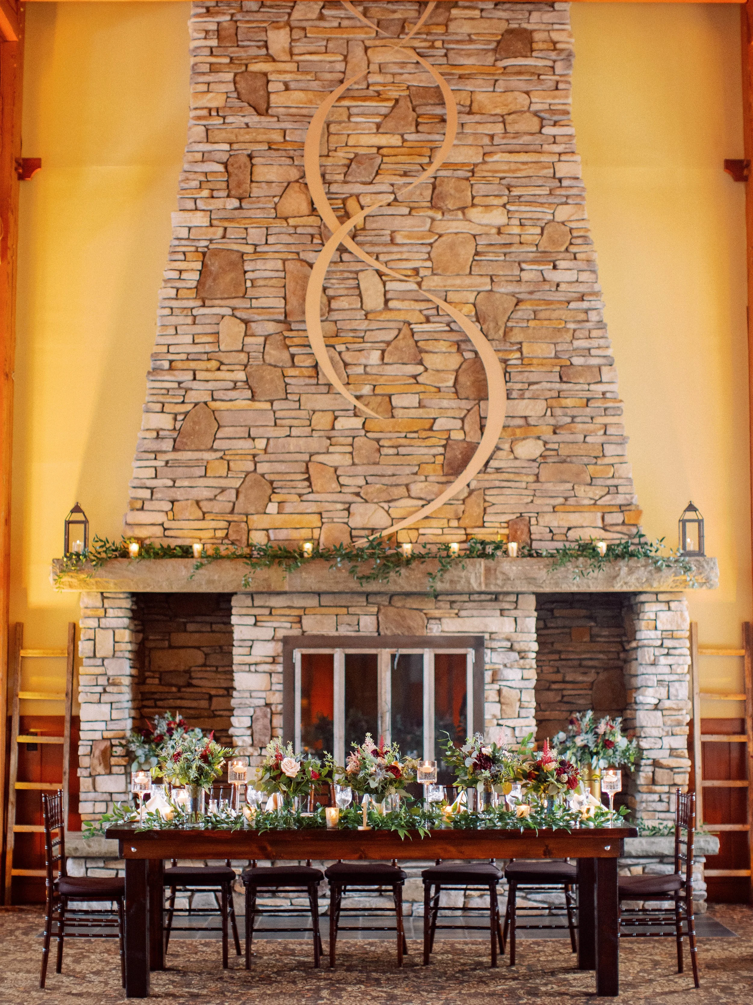 A decorated dining table set for an event in front of a large stone fireplace with a wall clock, candles, floral arrangements, and chairs.