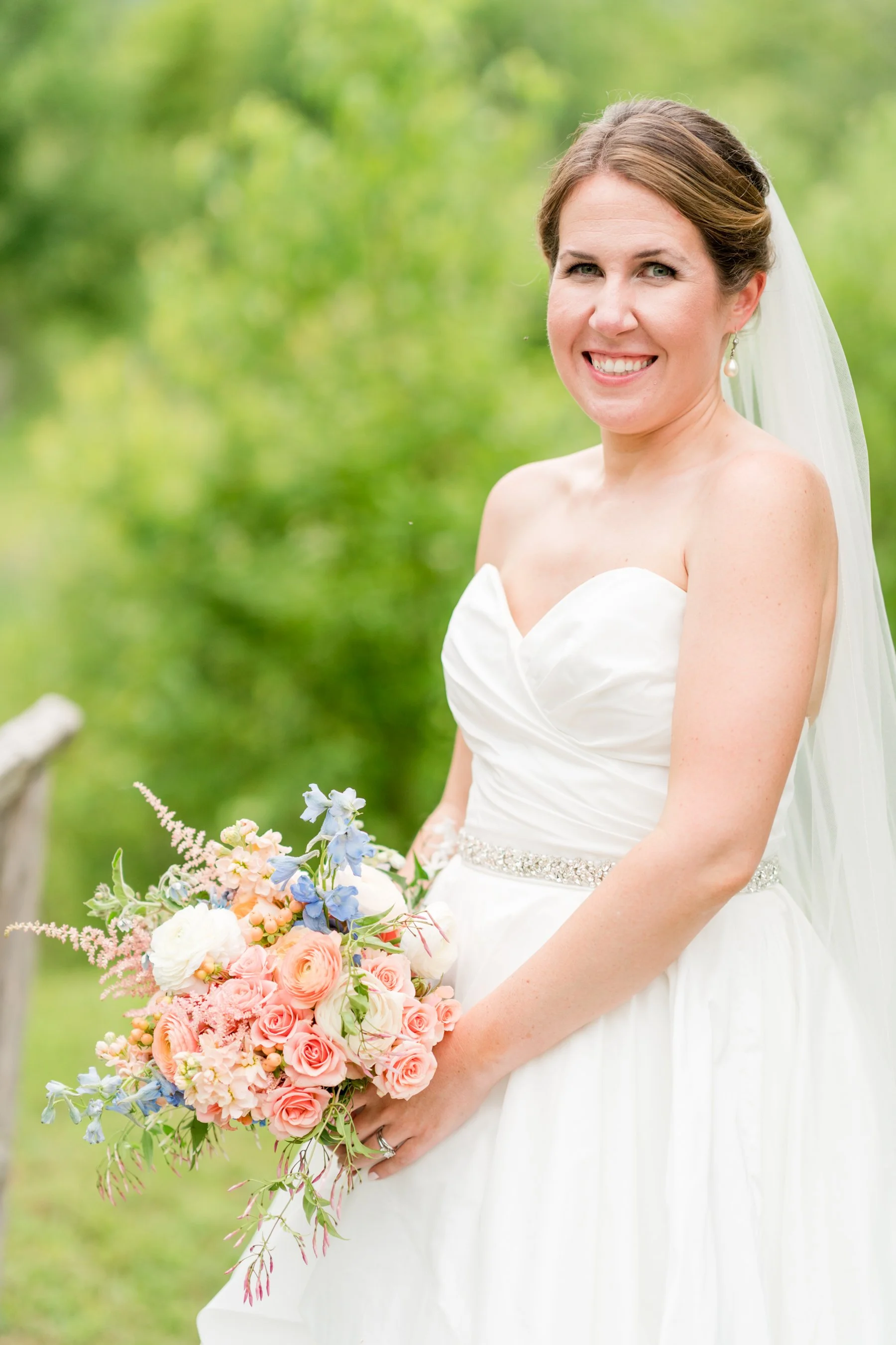 A woman smiling in a wedding dress, holding a large bouquet of pastel and colorful flowers, outdoors with a blurred green background.