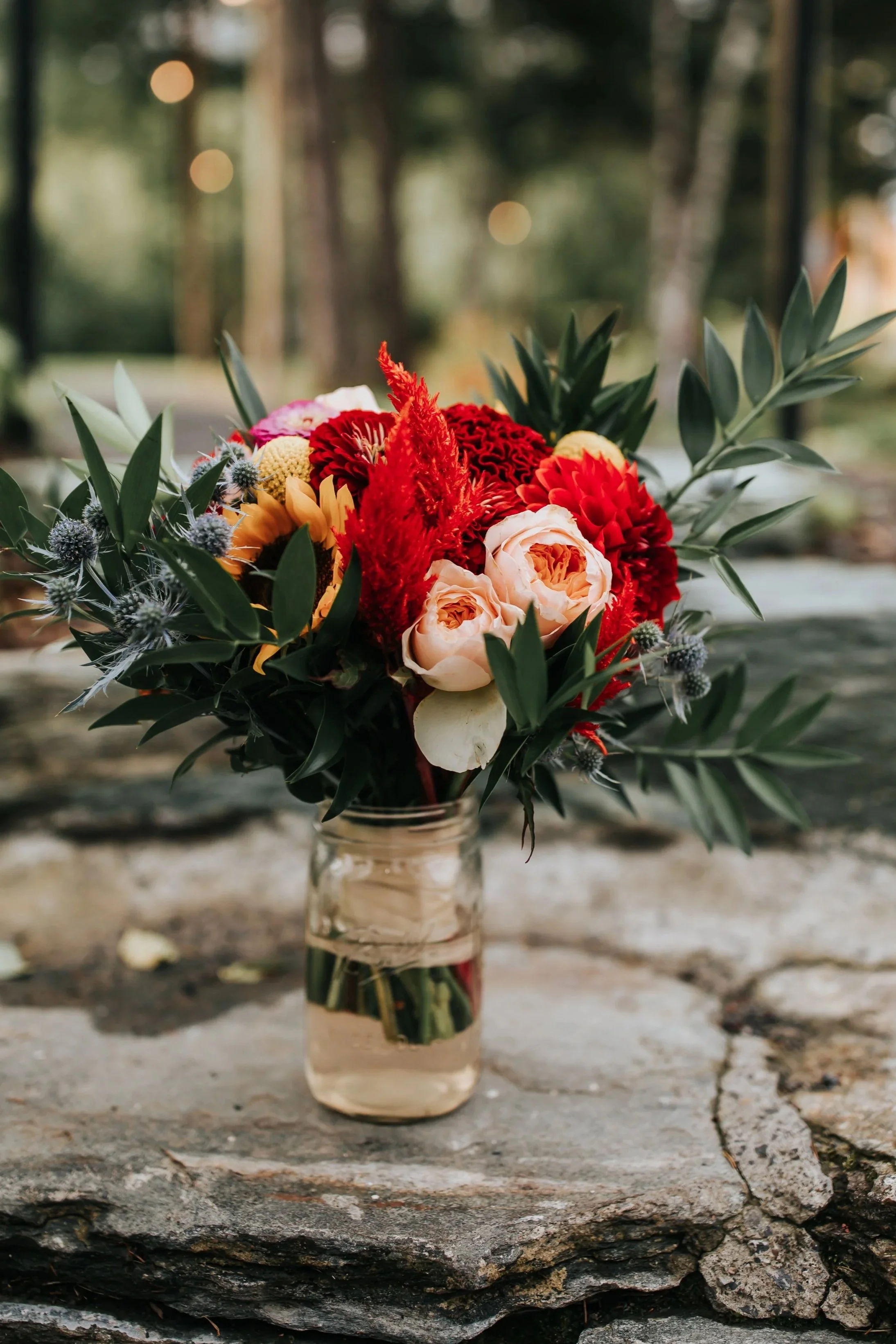 A bouquet of colorful flowers in a glass jar on a rustic wooden surface outdoors.