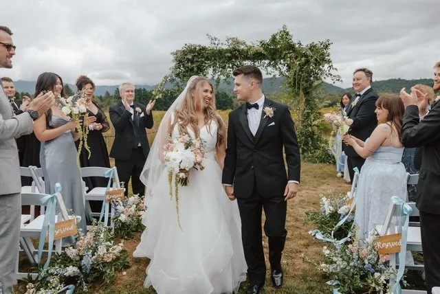 Bride and groom holding hands walking down the aisle during their wedding ceremony outdoors, with guests clapping and smiling, under a cloudy sky.