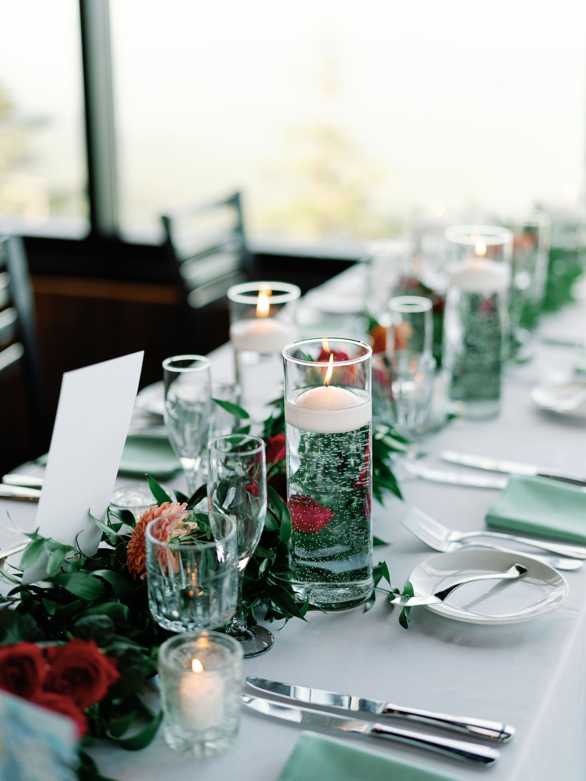 A table set for a special occasion with white plates, silverware, glasses, and candles inside a bright room, decorated with flowers and greenery.