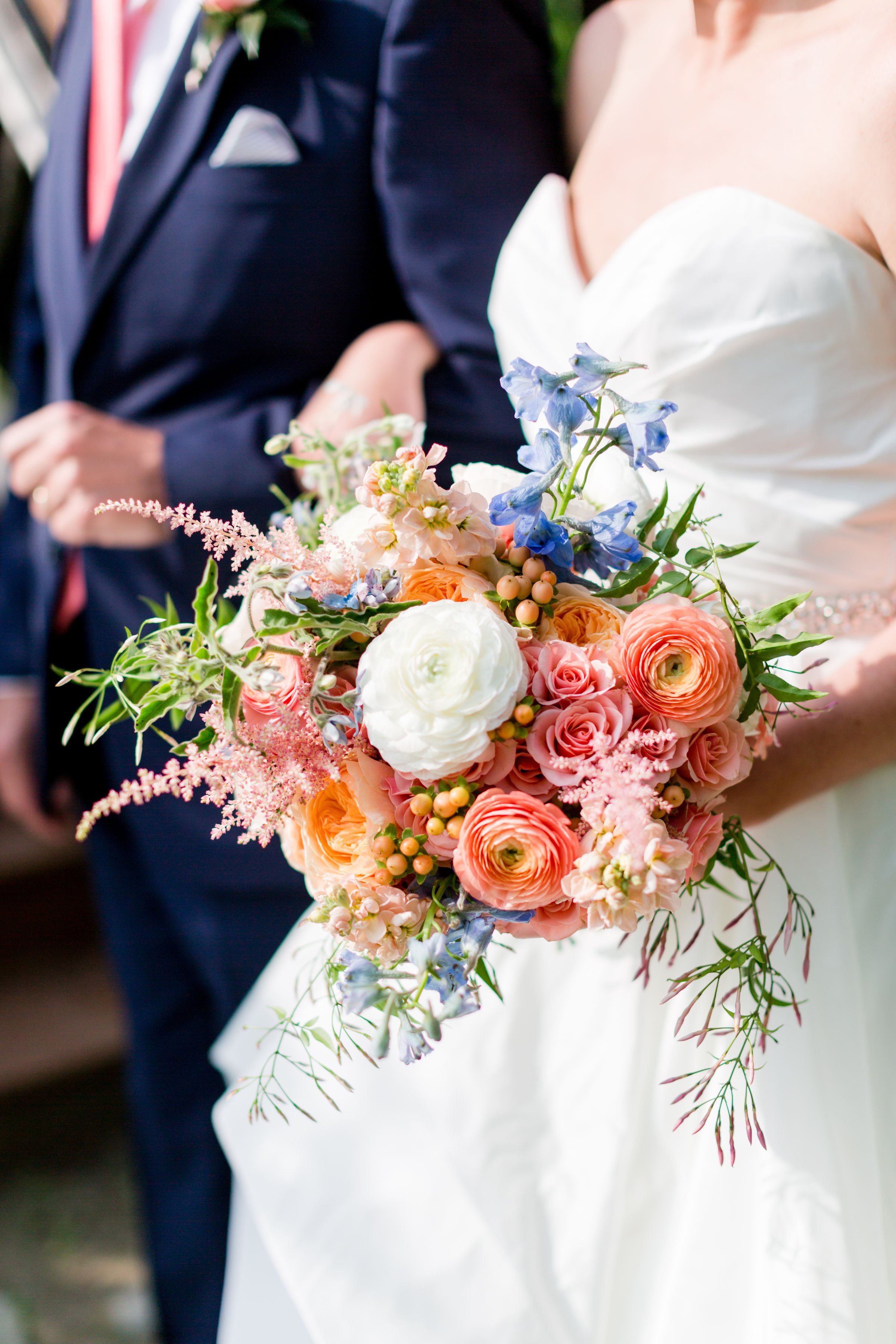 A bride holding a large bouquet of pink, white, peach, and blue flowers during a wedding ceremony with a groom in a dark navy suit beside her.