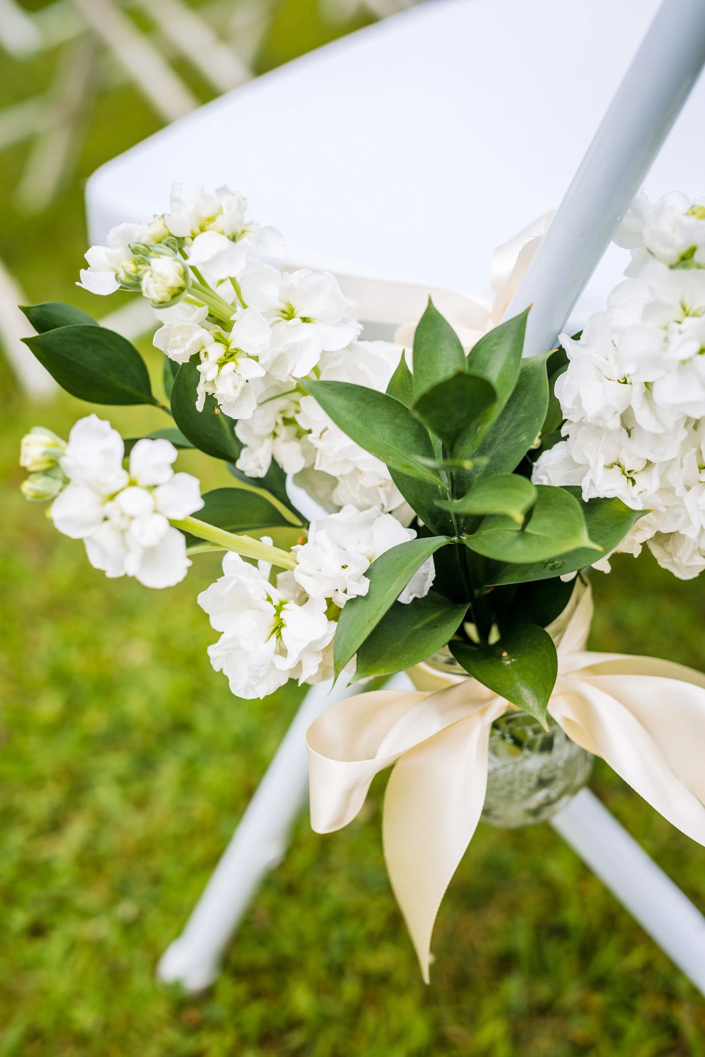 Close-up of white flowers with green leaves attached to a white seat post, with a cream-colored ribbon below, against a grassy background.