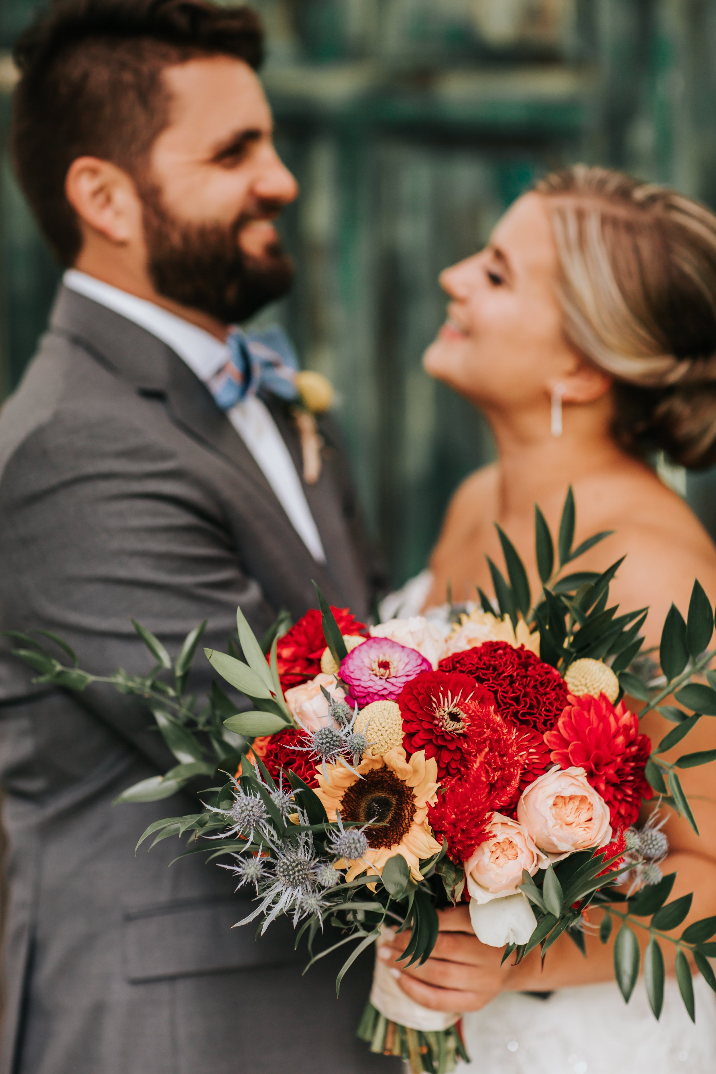 A bride holding a colorful bouquet of red, orange, pink, and white flowers with greenery, standing close to a groom dressed in a gray suit with a bow tie, during a wedding celebration.