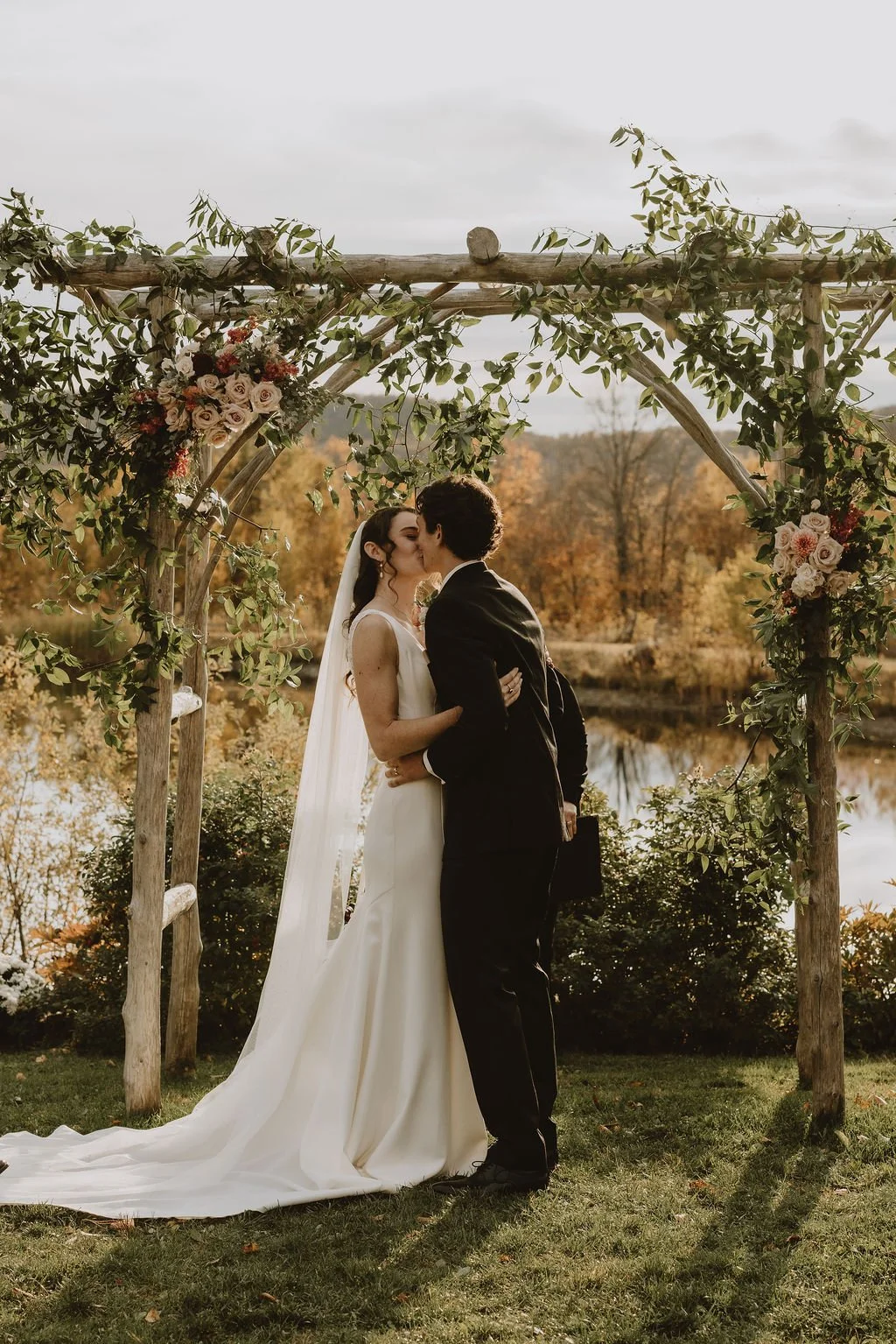 A bride and groom kiss under a wooden arch decorated with greenery and flowers outdoors, with a lake and autumn trees in the background.