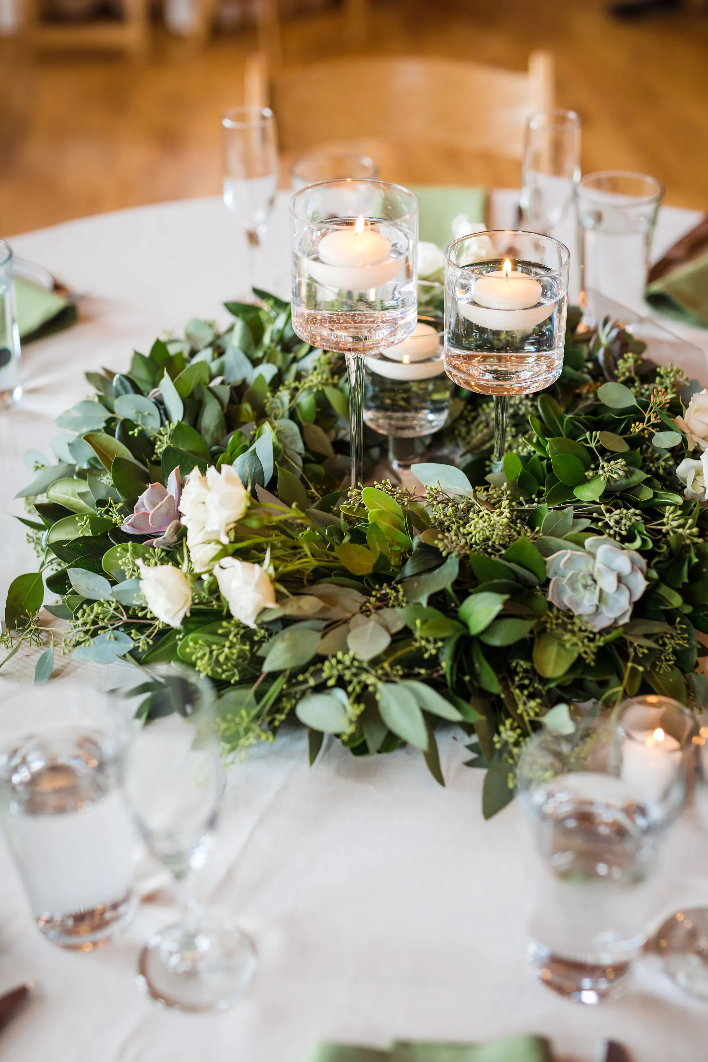 A round table decorated with a lush green and white floral centerpiece, surrounded by several glasses of water with floating candles, under warm ambient lighting.