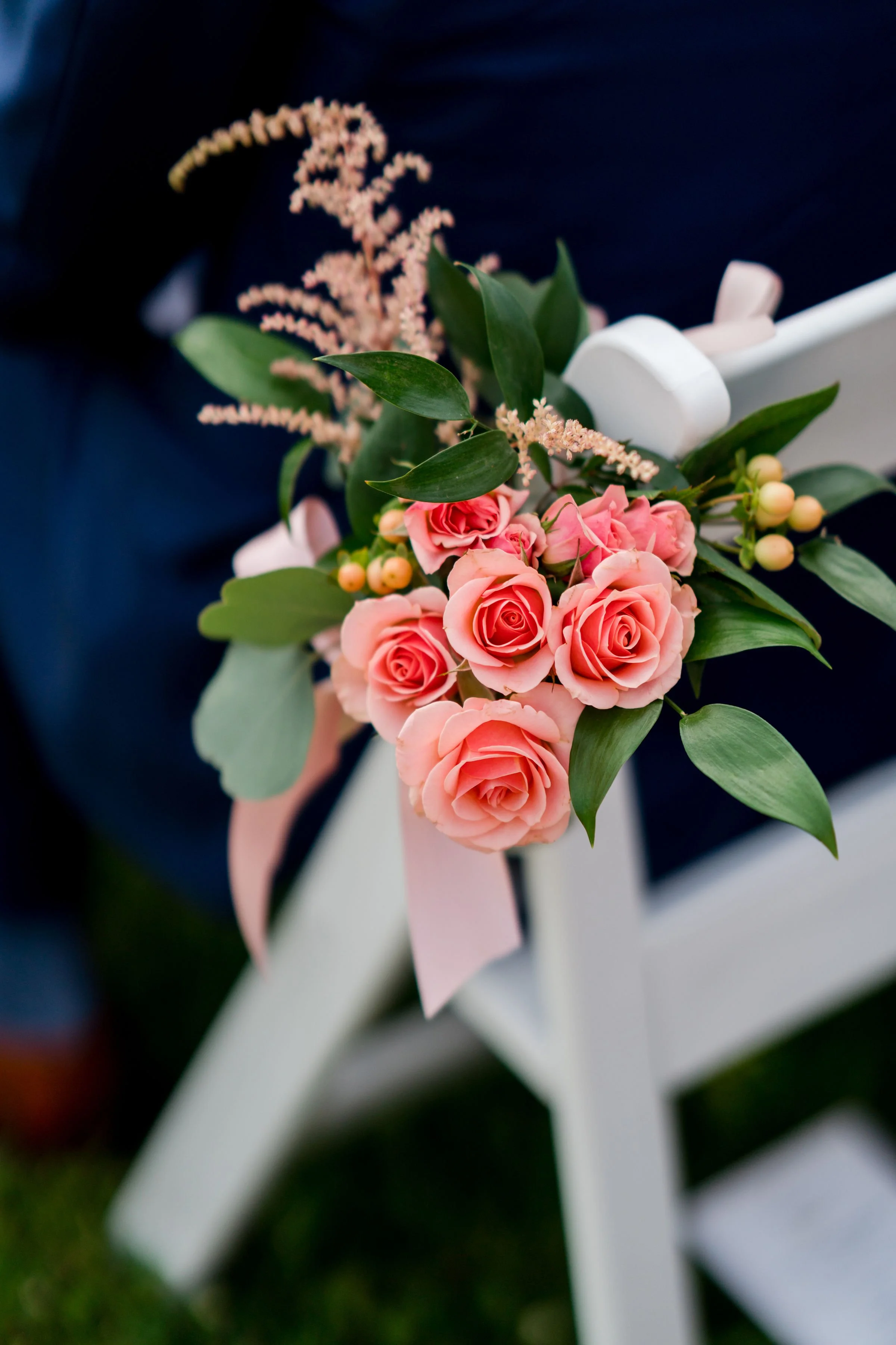 Close-up of a floral arrangement with pink roses, green leaves, and pink flowers against a dark background, attached to a white wooden chair with a pink ribbon.