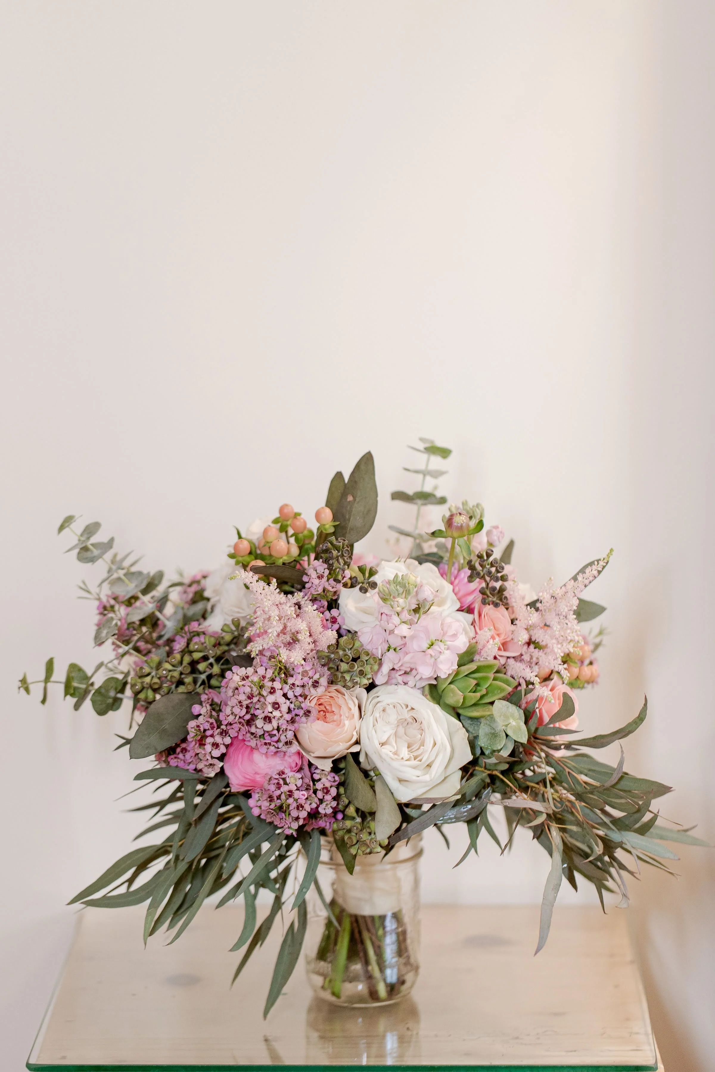 A large floral bouquet in a clear glass vase on a light-colored table against a plain off-white wall. The arrangement includes white roses, pink roses, pink and purple filler flowers, eucalyptus leaves, and other greenery.