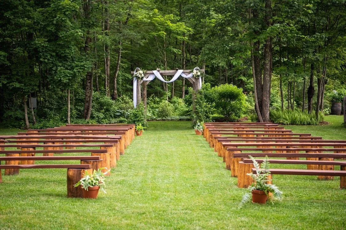 Empty outdoor wedding ceremony setup with wooden benches on either side of a grassy aisle, leading to a decorated wooden arch in a wooded area.