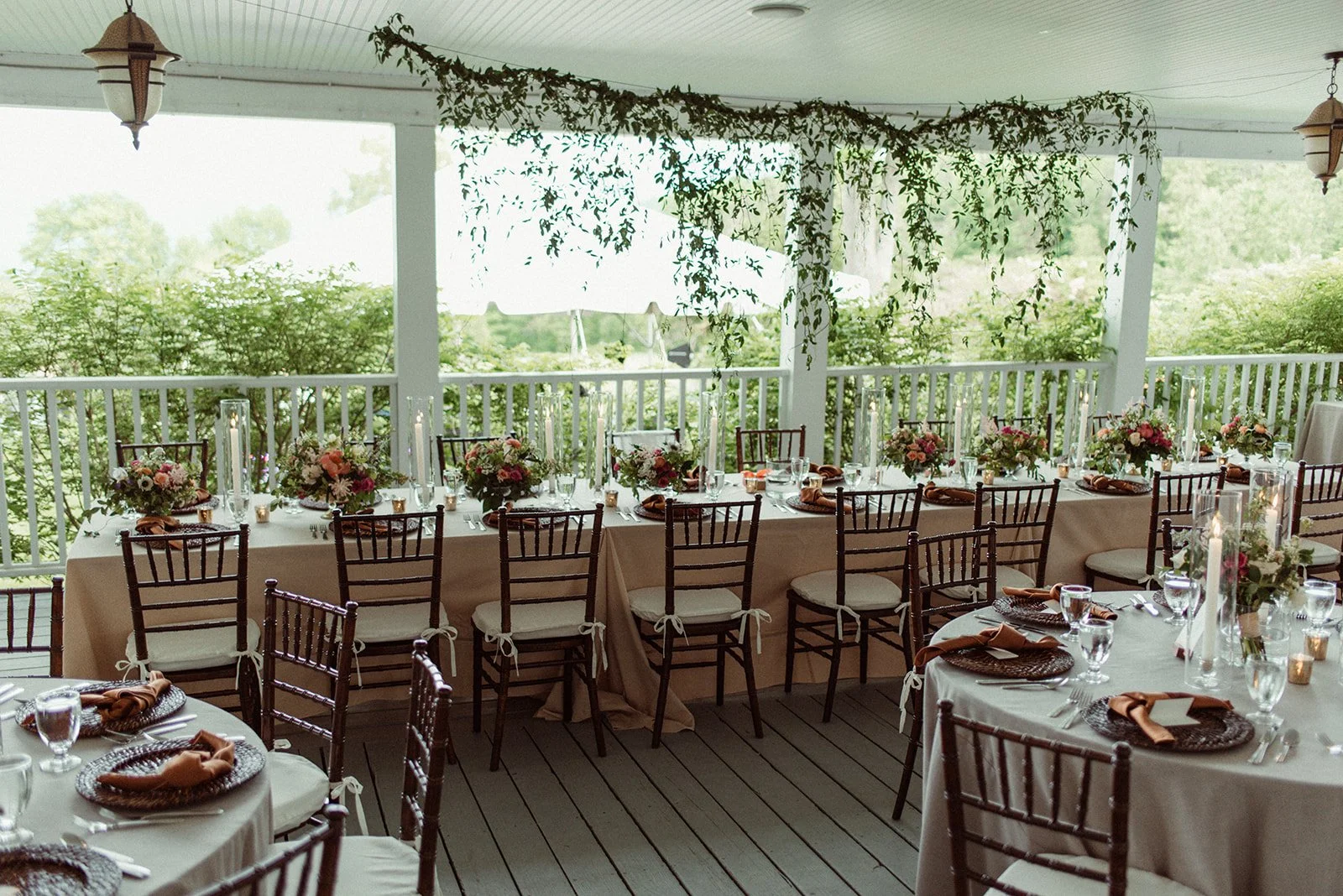 A decorated outdoor dining area with round and rectangular tables covered in white tablecloths, set with glasses, silverware, napkins, and brown chargers. The tables are adorned with floral centerpieces and tall candles, and surrounded by wooden chai