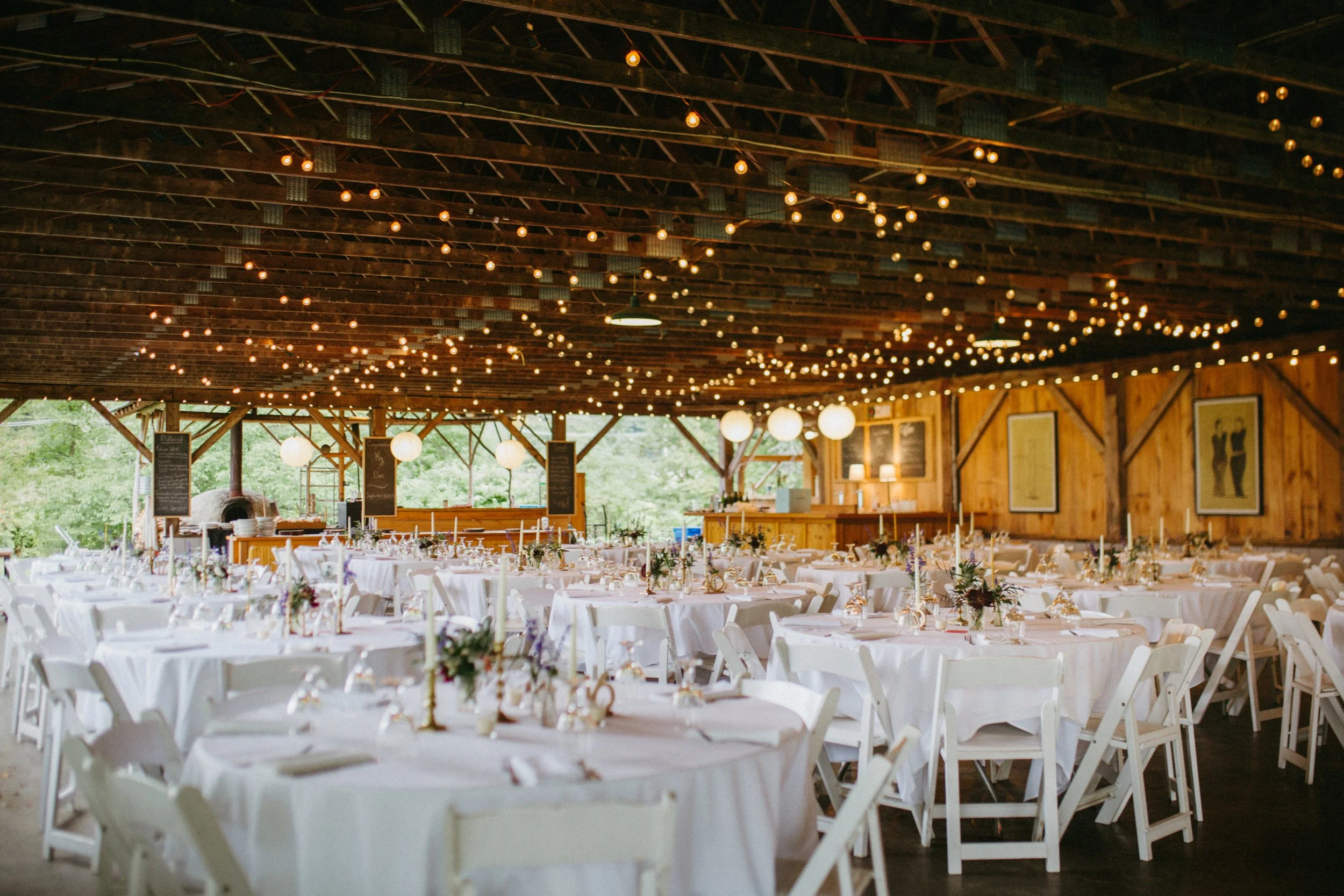 Wedding reception area with round tables covered in white tablecloths, decorated with flowers and candles. The rustic barn interior features a wooden ceiling with string lights hanging down, creating a warm ambiance. Large windows and framed artwork 