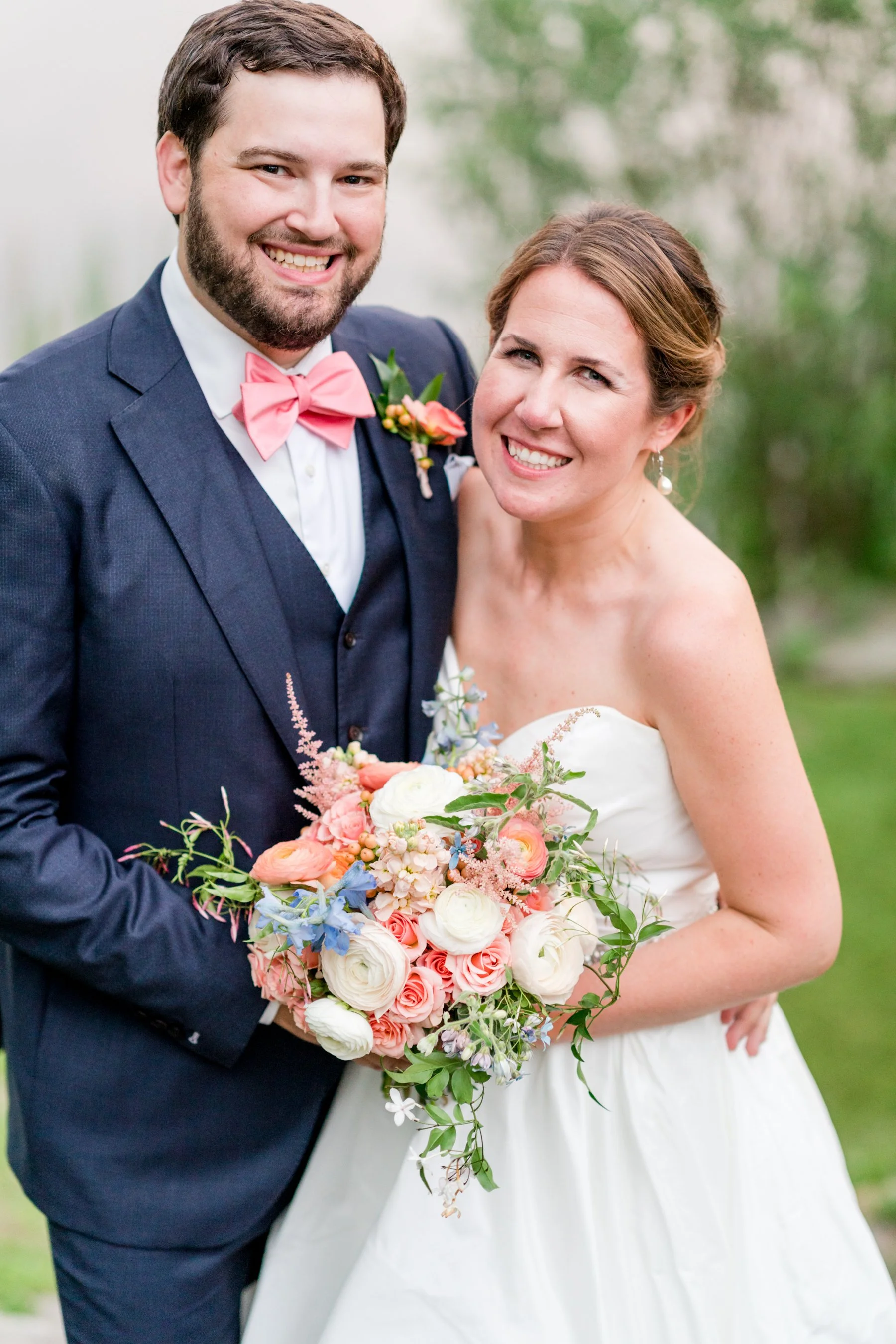 A smiling wedding couple outdoors with a blurred green background. The groom in a navy suit with a pink bow tie and boutonniere holds a peony, and the bride in a strapless white gown holds a colorful floral bouquet.