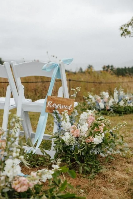 White chair with light blue ribbon, reserved sign, and floral arrangements on grass outdoors, overcast sky in the background.