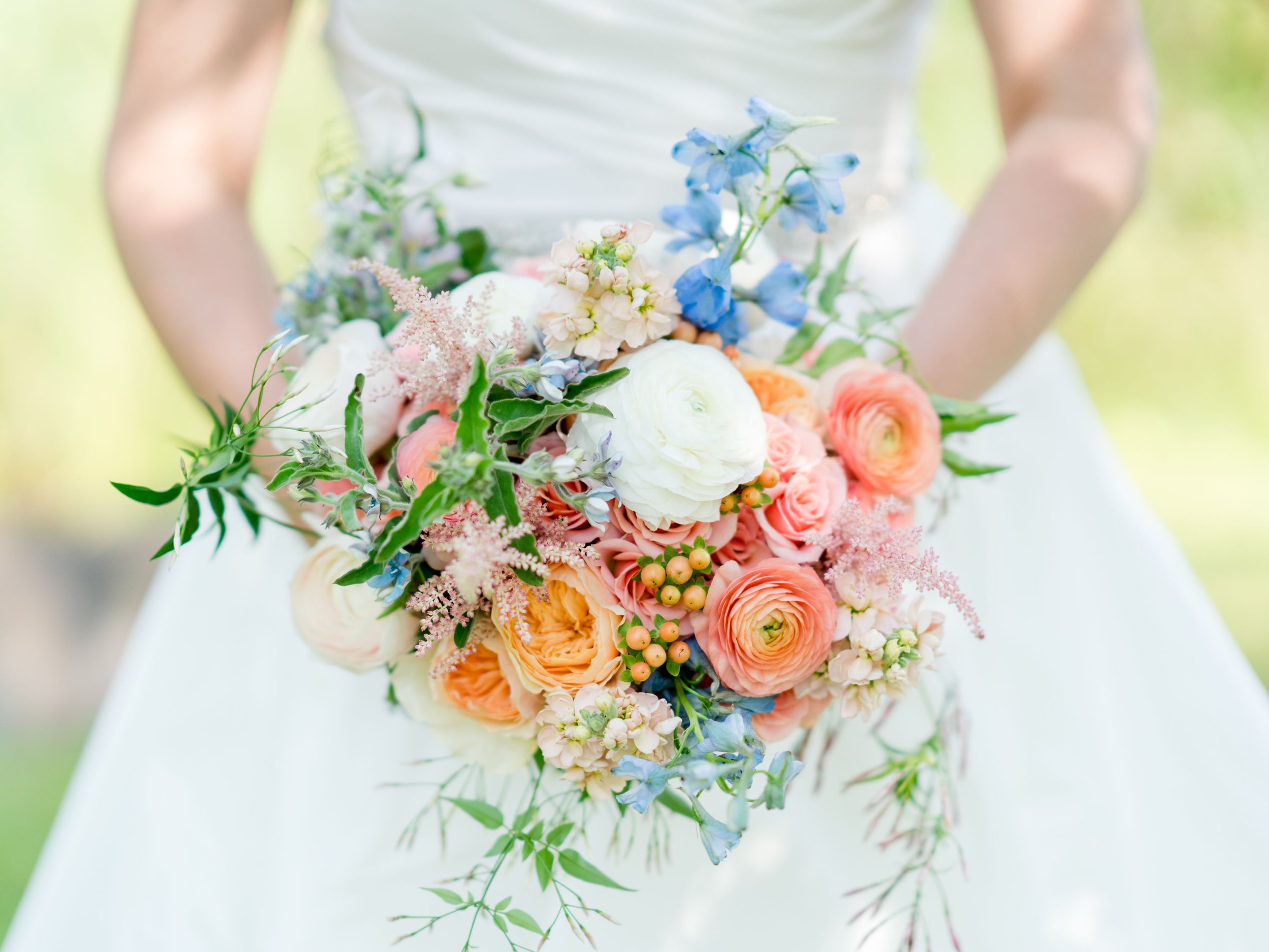 Close-up of a person holding a bouquet of colorful flowers, including white, pink, peach, and blue blossoms, against a blurred outdoor background.