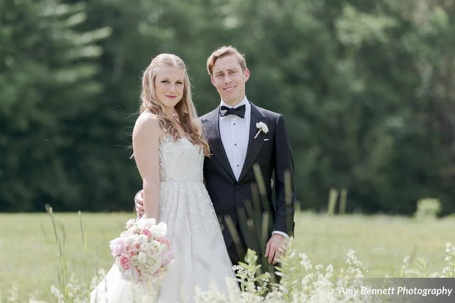 A bride and groom standing outdoors in a grassy field, with trees in the background, smiling at the camera during their wedding.