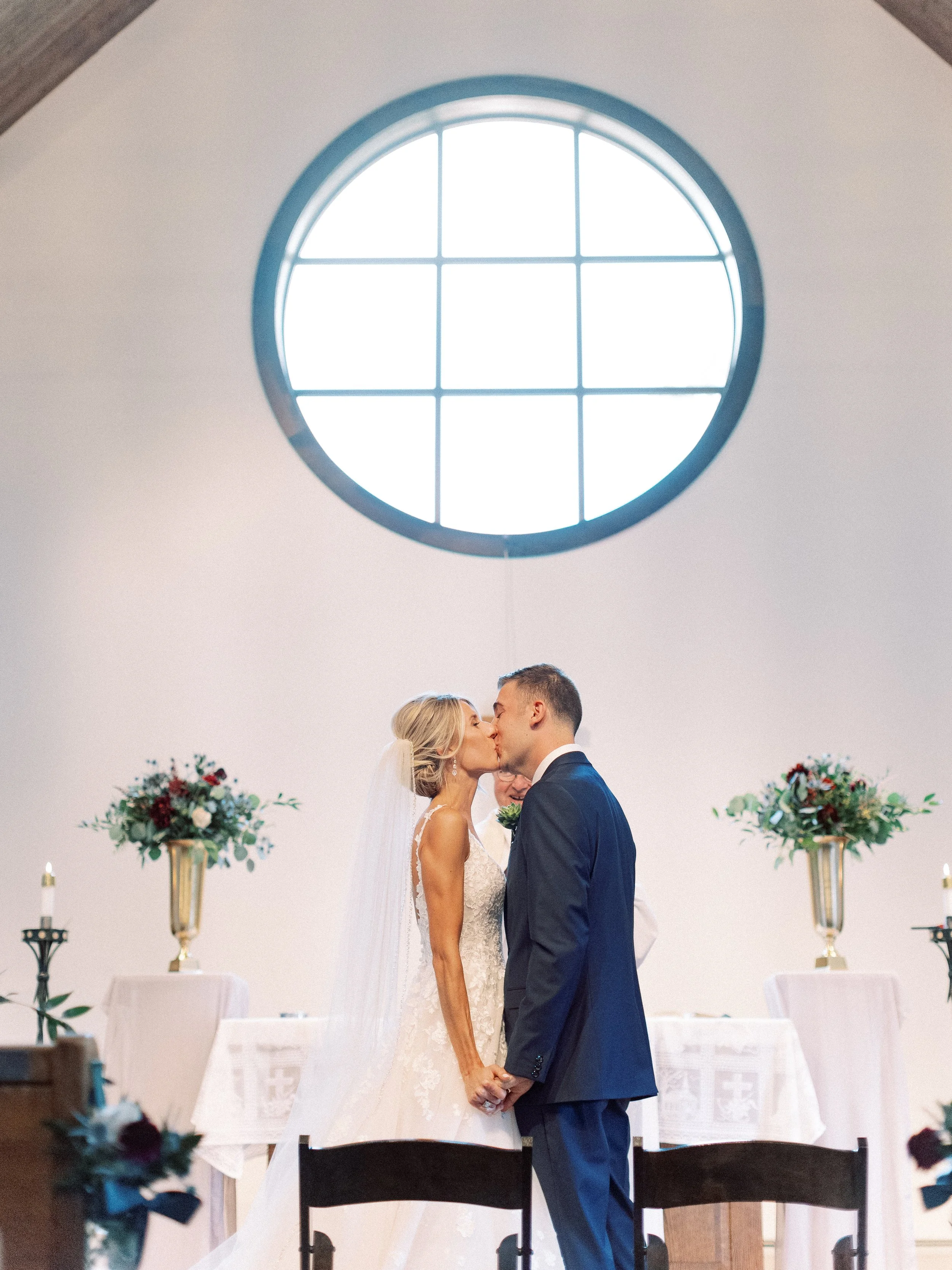 A bride and groom kiss during their wedding ceremony inside a church or chapel with a large circular window in the background, and floral arrangements on tall stands on either side.