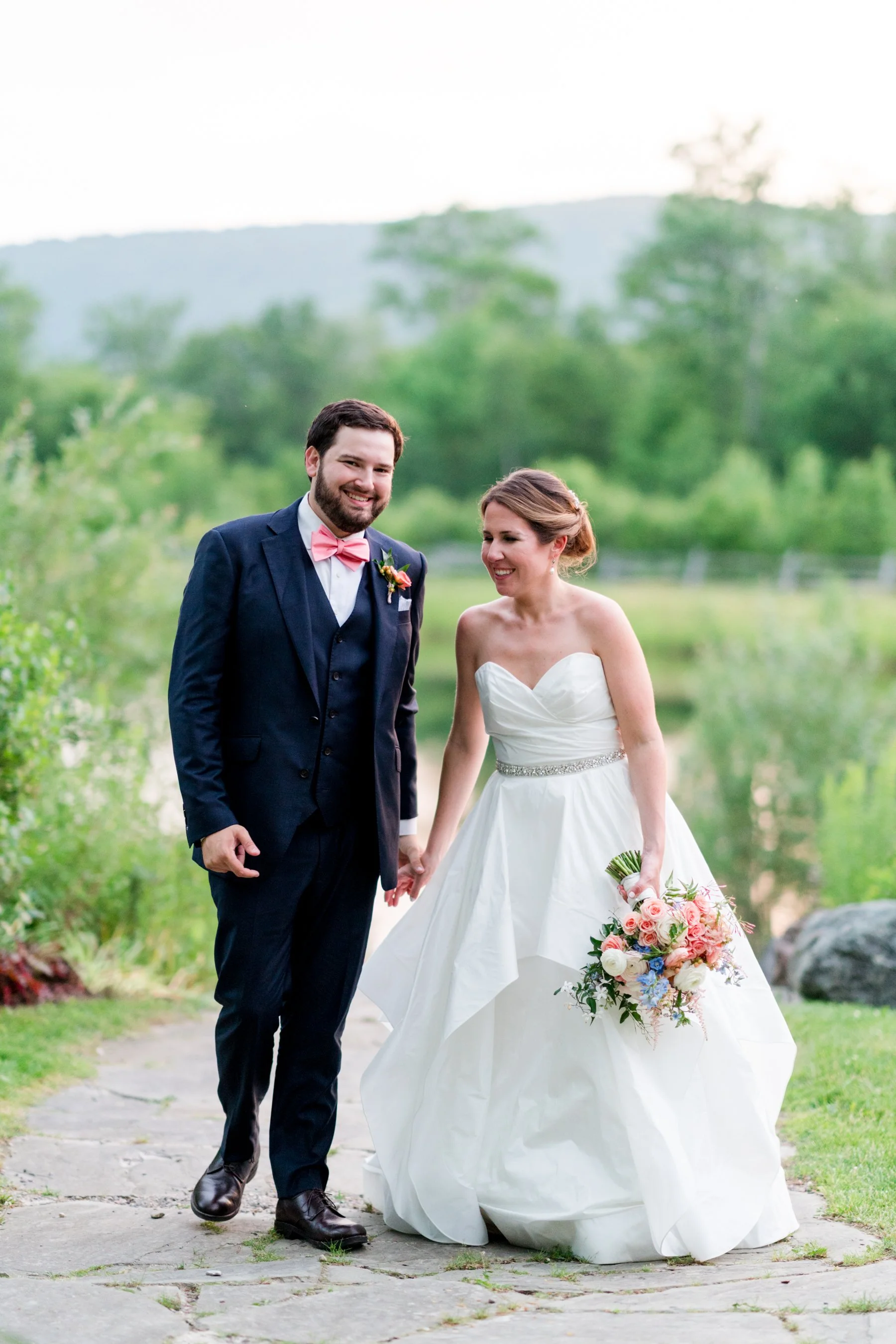 A newlywed couple holding hands and smiling outdoors, with a green landscape and trees in the background. The groom is dressed in a navy suit with a pink bow tie, and the bride is in a white strapless wedding gown holding a bouquet of flowers.