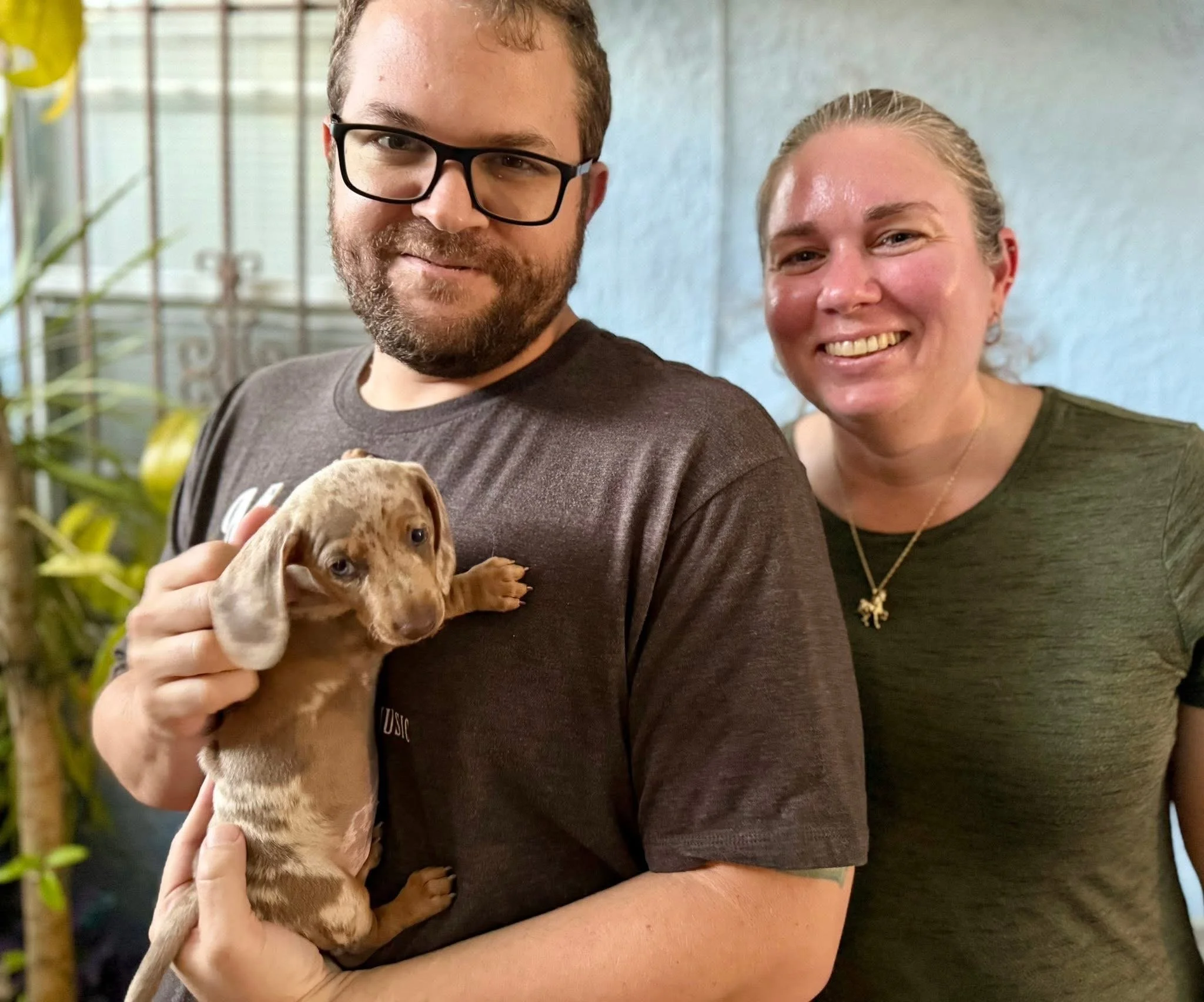 Two smiling people, a man with glasses and a woman, holding a small brown puppy with floppy ears.