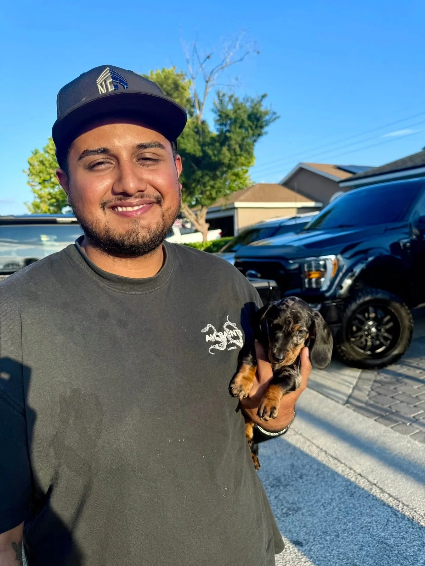 A man with a beard and mustache smiling while holding a small black and brown puppy, standing outside on a sunny day with houses, trees, and parked cars in the background.