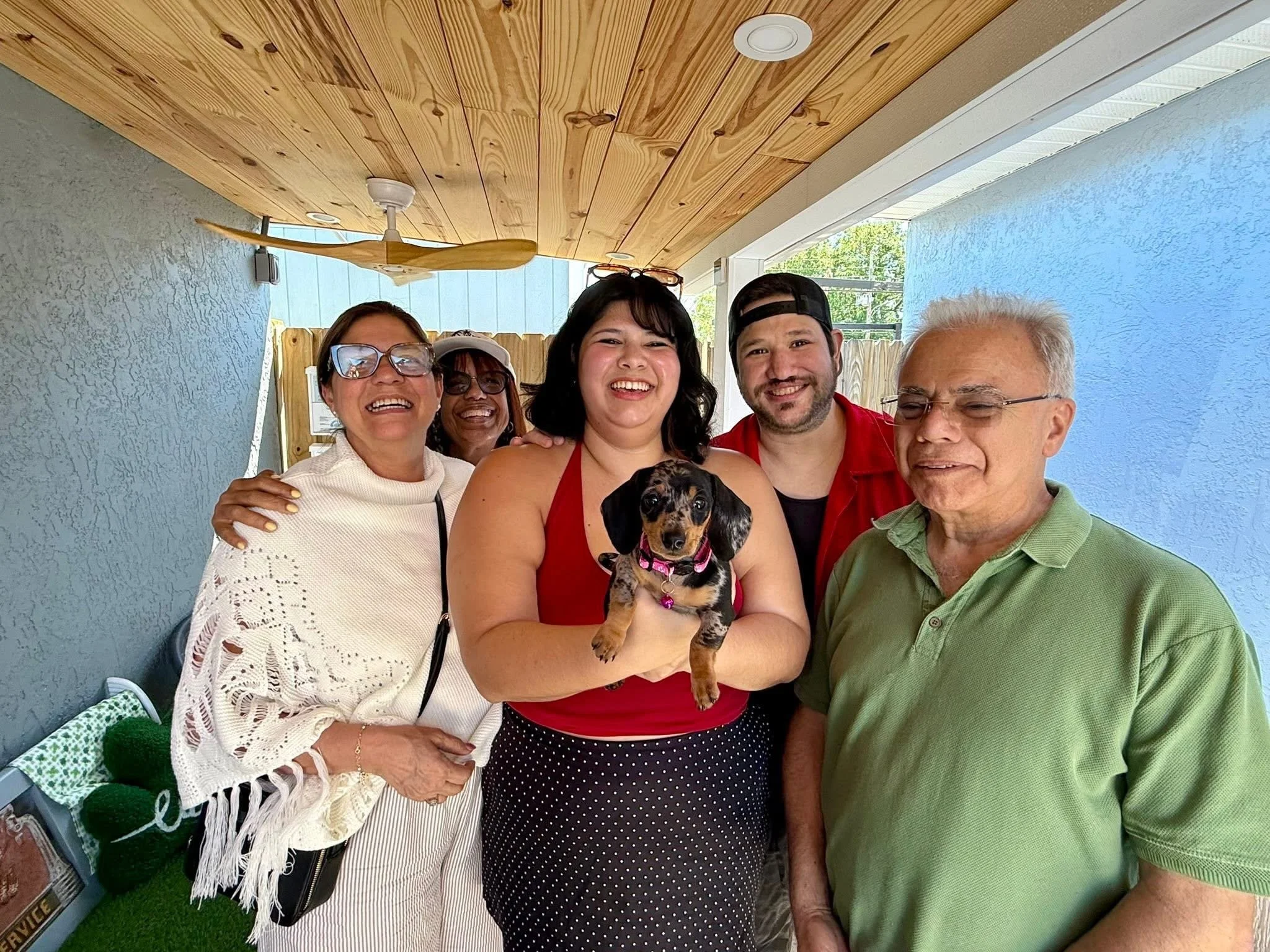 Group of five people, four women and one man, standing on a porch, smiling with a puppy. An elderly woman in the front left wears glasses and a white crochet top, the woman behind her has dark hair and glasses, the woman in the center has short dark 