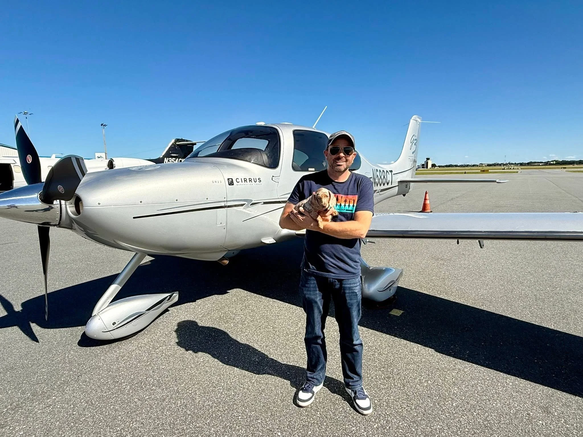 A man wearing sunglasses, a cap, and sneakers holding a pug dog in front of a small silver airplane on an airport runway under a clear blue sky.