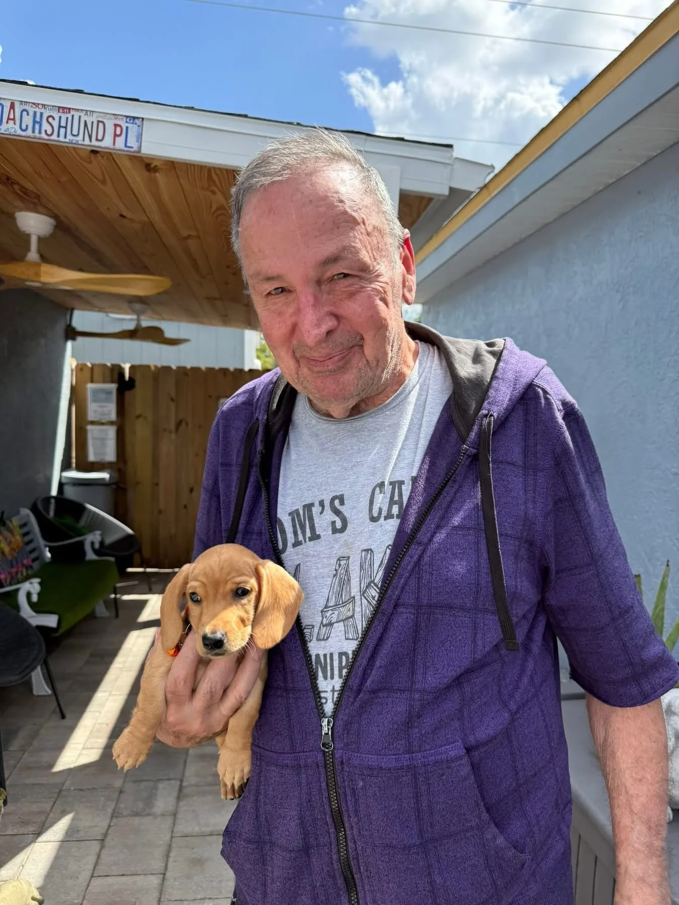 An older man with gray hair holding a small tan puppy puppy, standing outside in a backyard with a wooden fence, garden chairs, and a house. The man is smiling and wearing a gray T-shirt and a purple jacket.