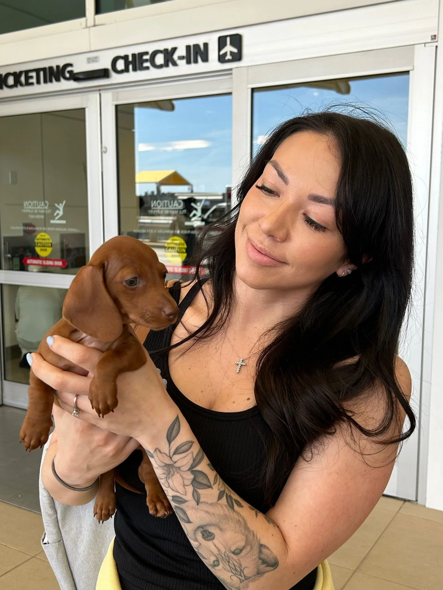 A woman with dark hair and tattoos on her arm holding a brown puppy at an airport check-in area.