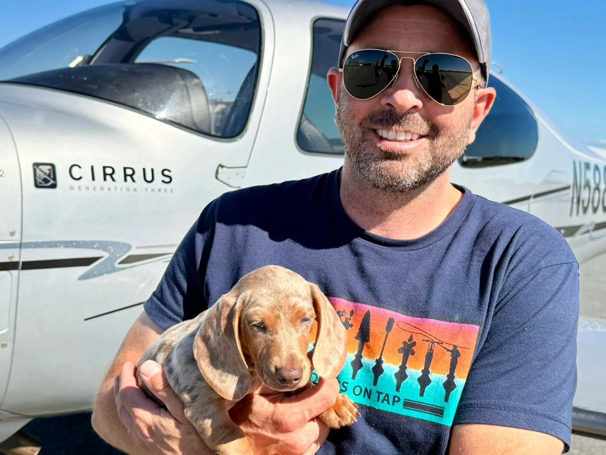 A man wearing sunglasses and a cap holding a sleeping puppy with tan fur and black spots, standing in front of a small aircraft with the logo 'CIRRUS' on the side.