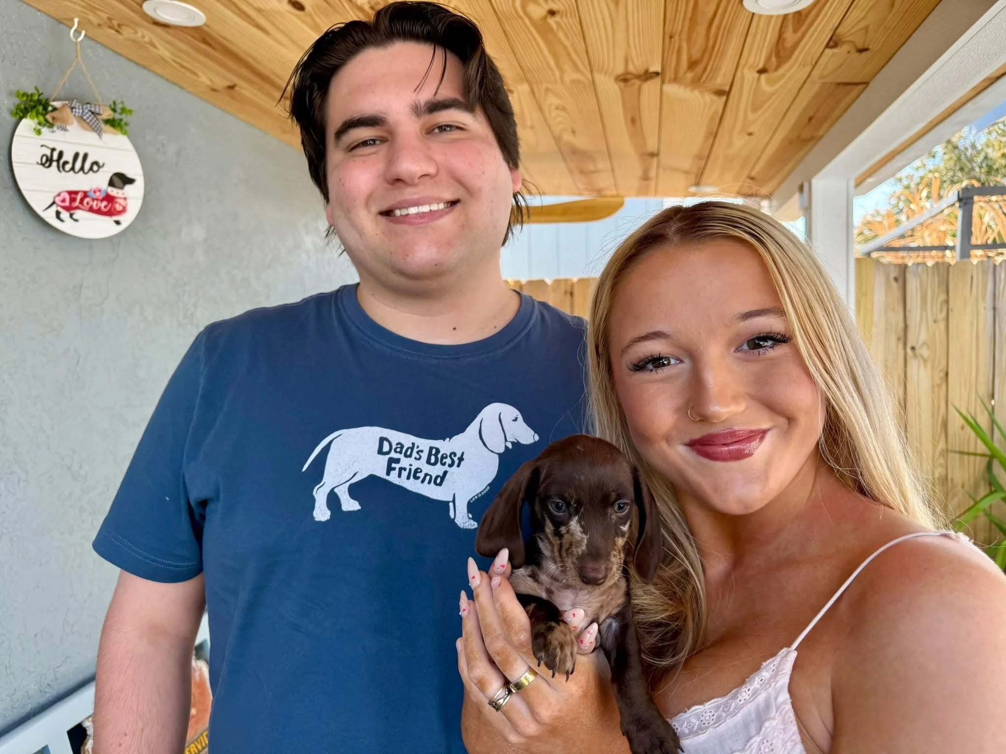 A young woman with blonde hair and a septum piercing holds a small, brown and black puppy with floppy ears, a nose, and wide eyes. Behind her, a man with dark hair, wearing a blue t-shirt, smiling. The background features a wall with a decorative sig