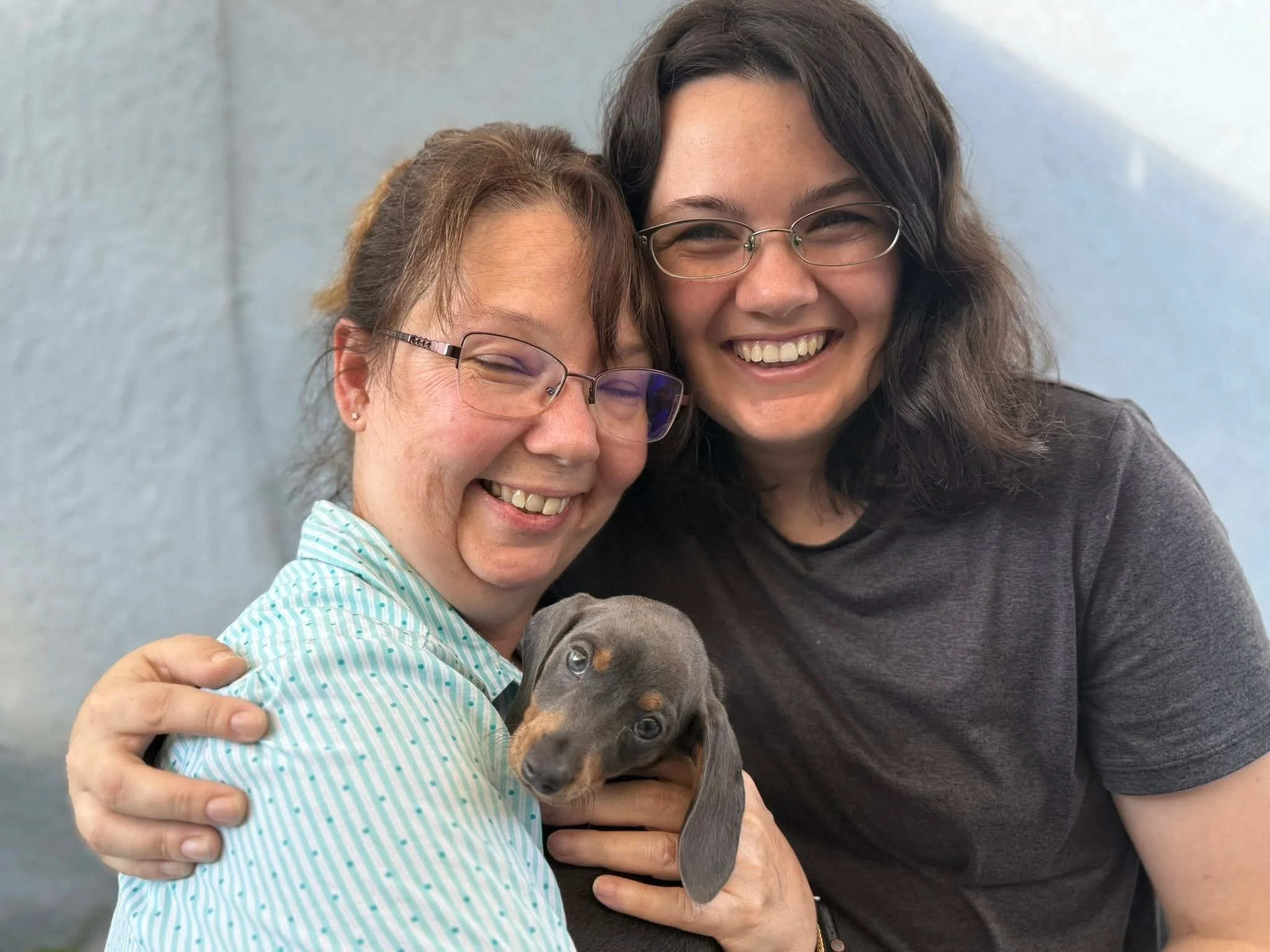 Two women hugging and smiling with a puppy in front of a plain light-colored wall.