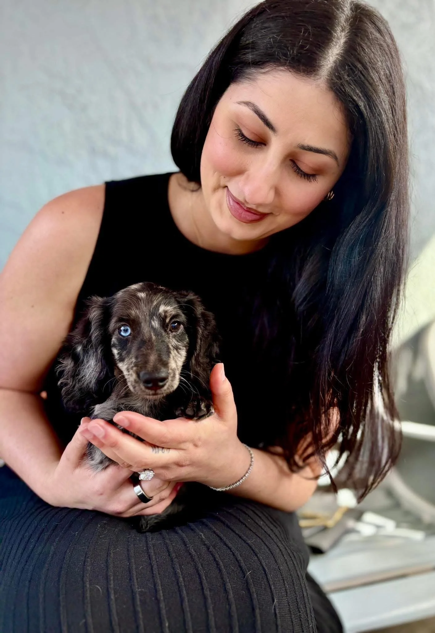 A woman with long dark hair, wearing a black sleeveless top, holding a black and gray puppy with blue eyes, sitting indoors with a light-colored wall in the background.