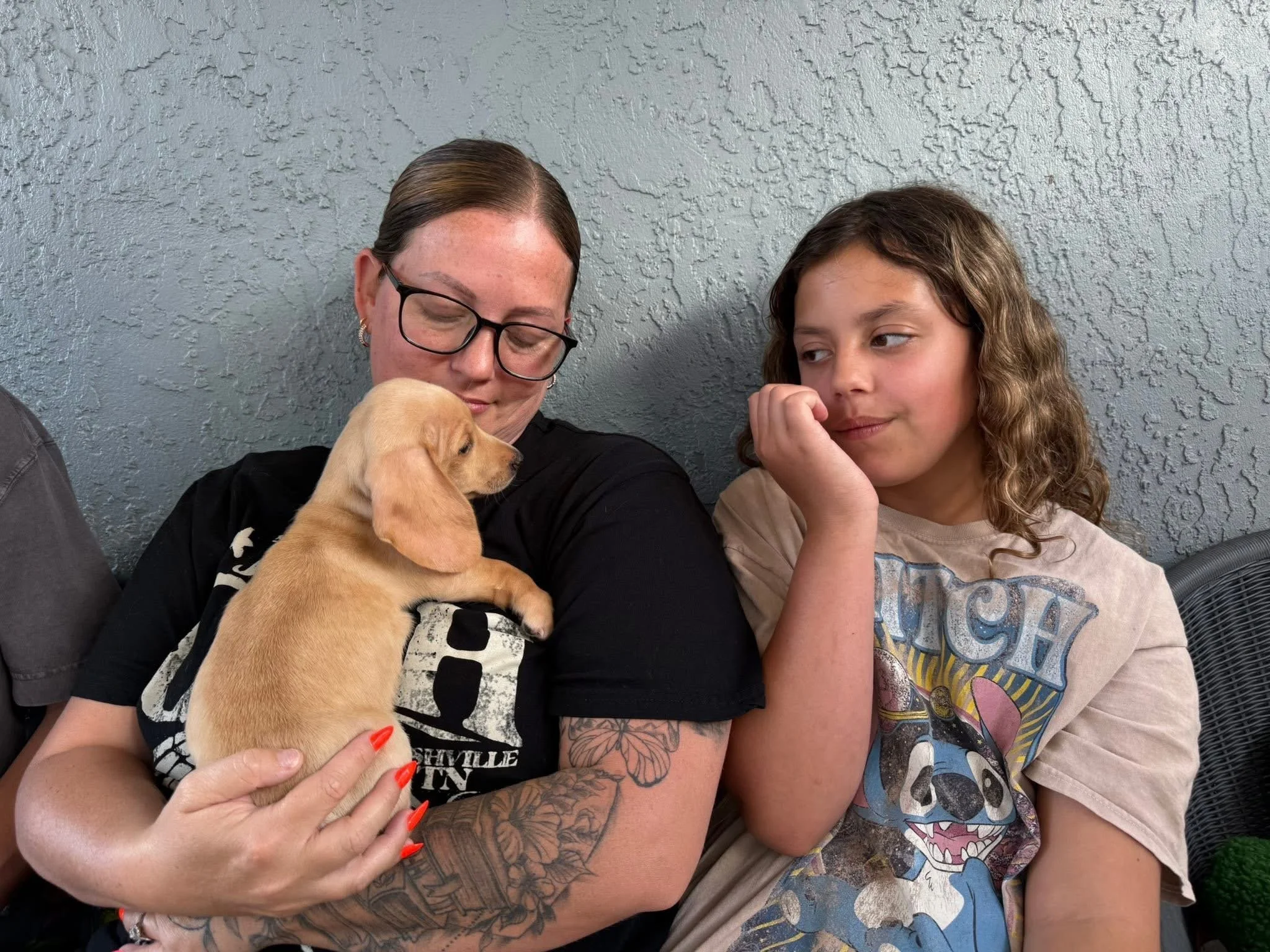 A woman with glasses and tattoos on her arm is holding a light-colored puppy. A young girl with curly hair is sitting nearby, resting her chin on her hand and looking at the puppy.
