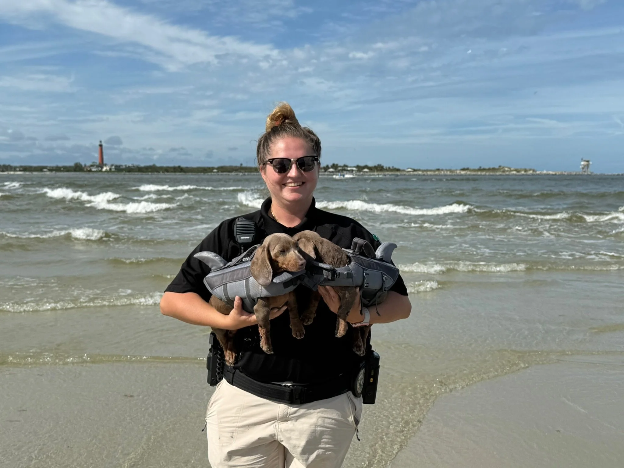 Woman at the beach holding three puppies in a pet carrier with ocean waves behind her.