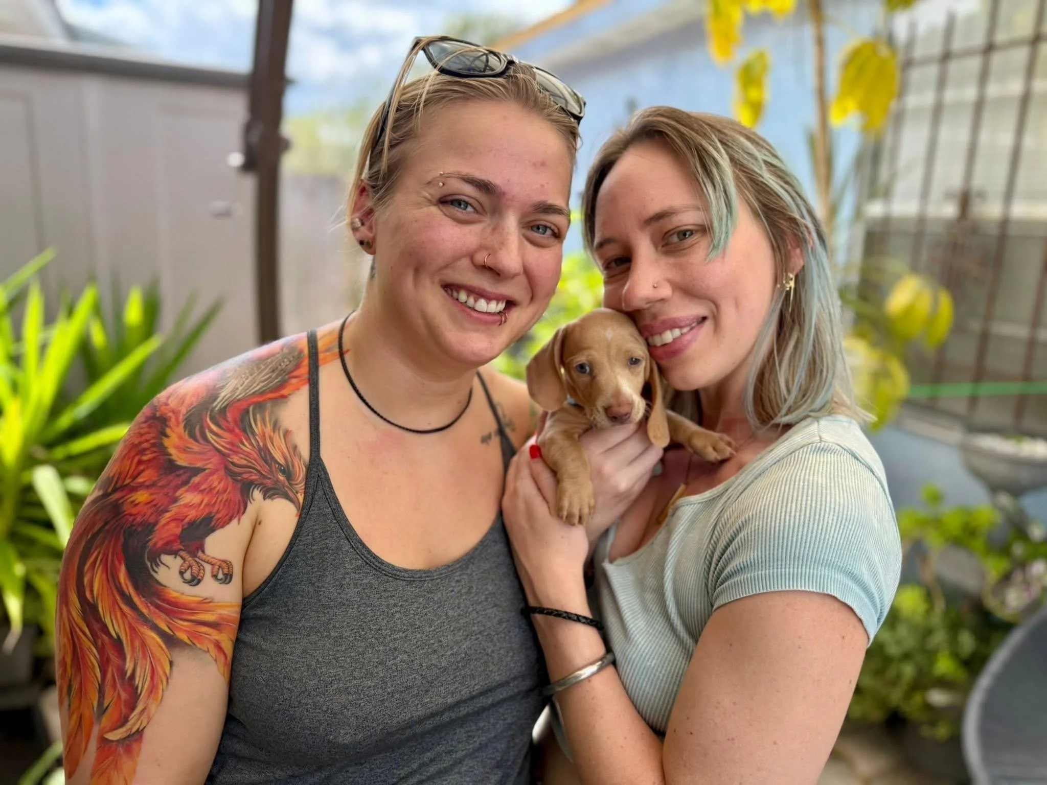 Two women smiling and holding a small brown puppy outdoors, with greenery and a greenhouse-like structure in the background.