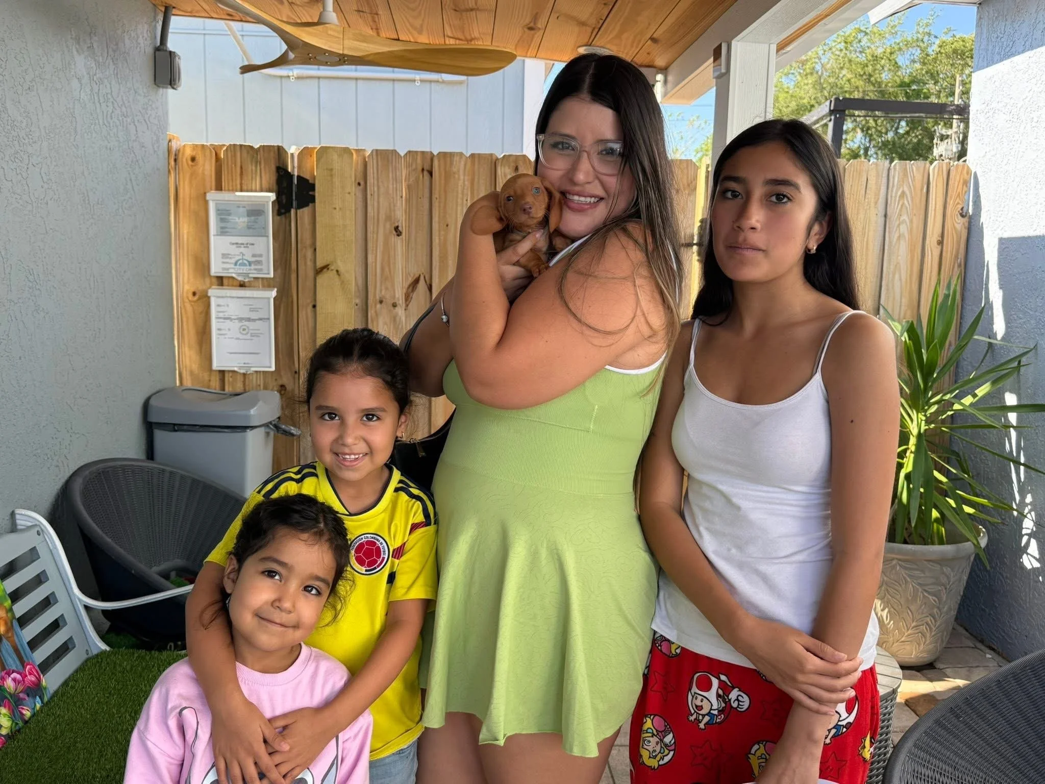 A group of children and two women standing outdoors in a backyard, with some holding a small puppy. The scene is bright and sunny with trees in the background.