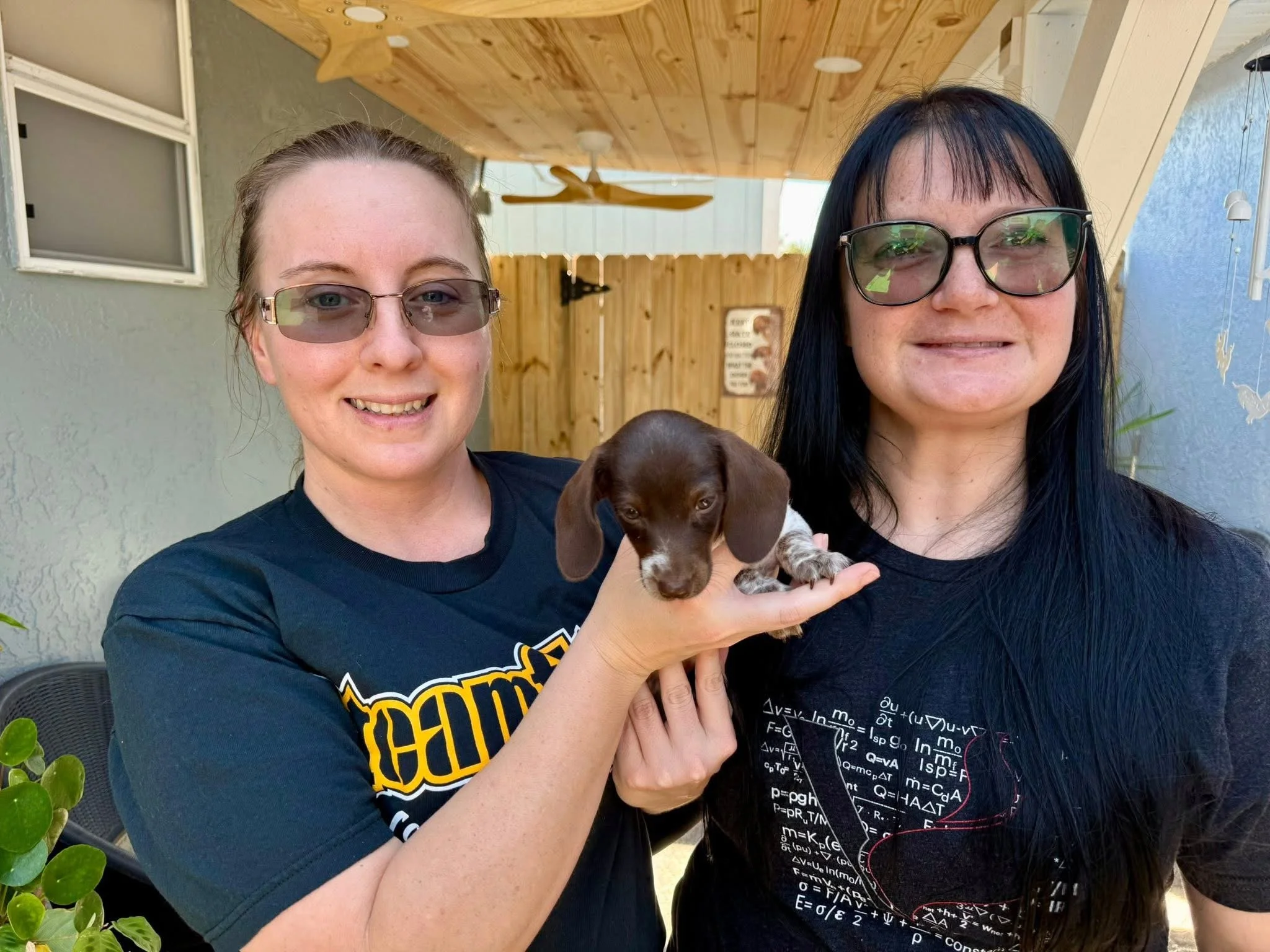 Two women wearing glasses holding a dachshund puppy outdoors, with a wooden ceiling, ceiling fan, and a wooden fence in the background.