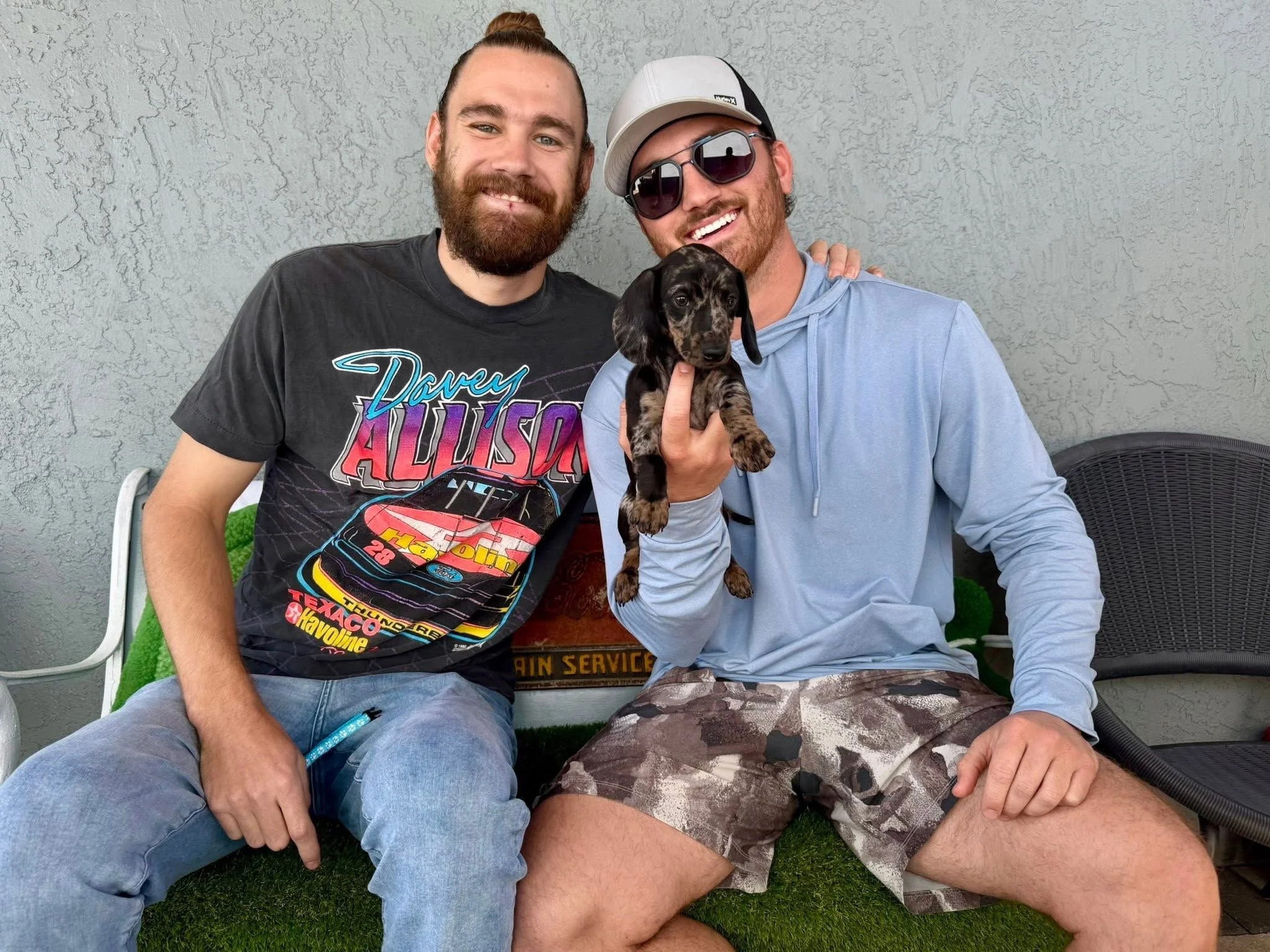 Two men sitting outdoors against a textured gray wall, smiling, one holding a small black and brown puppy, dressed casually in T-shirts, shorts, and sunglasses.