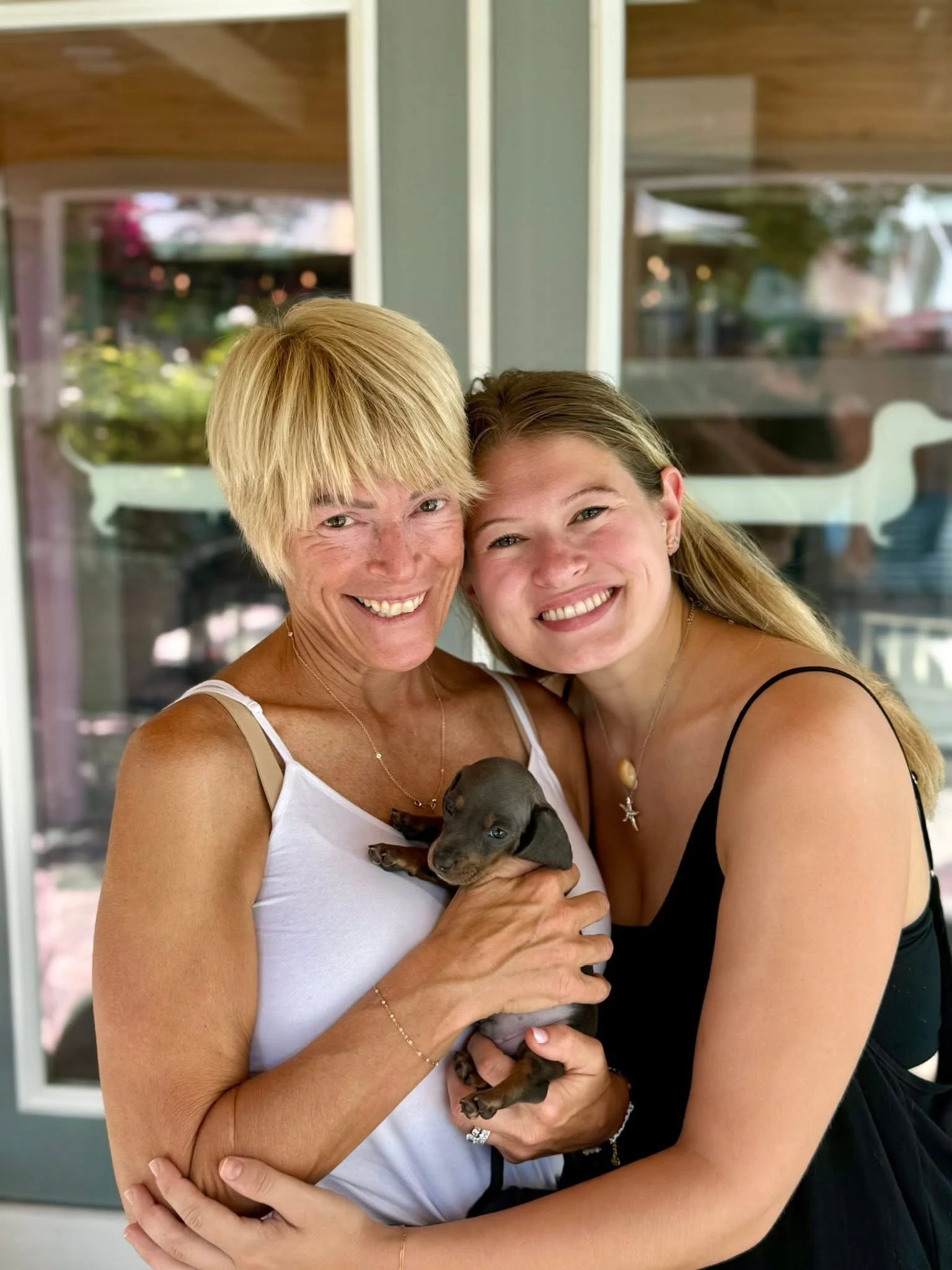 Two women hugging and smiling, holding a black puppy with a face and paws visible.