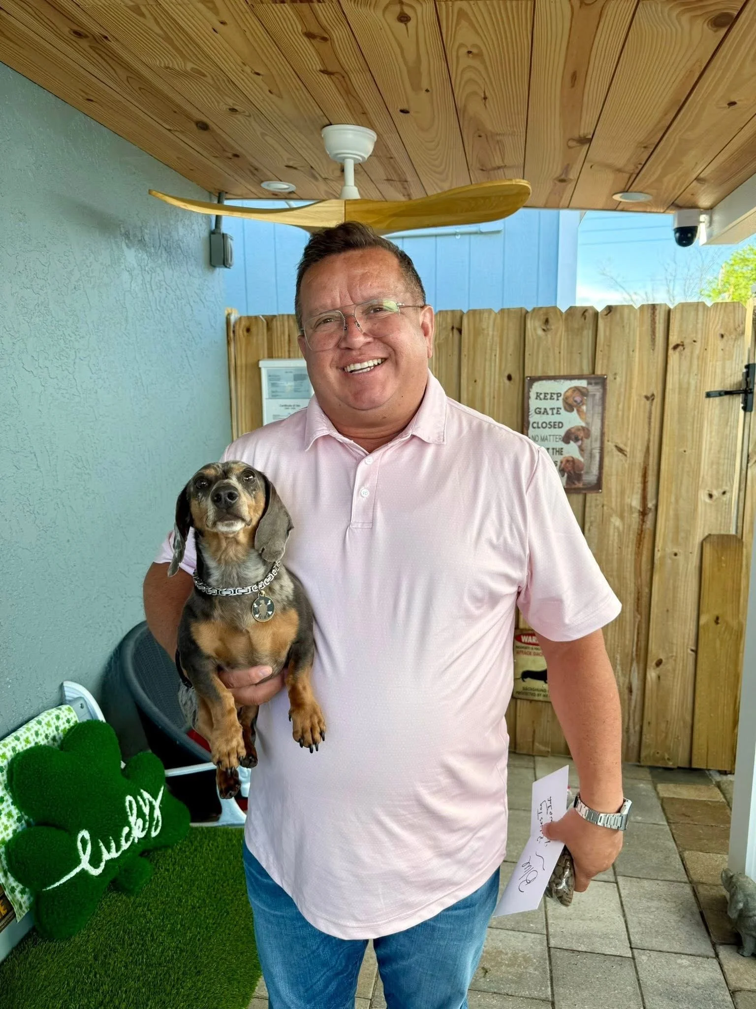 A man smiling and holding a small dog with floppy ears, standing near a wooden fence with a sign, during daytime.