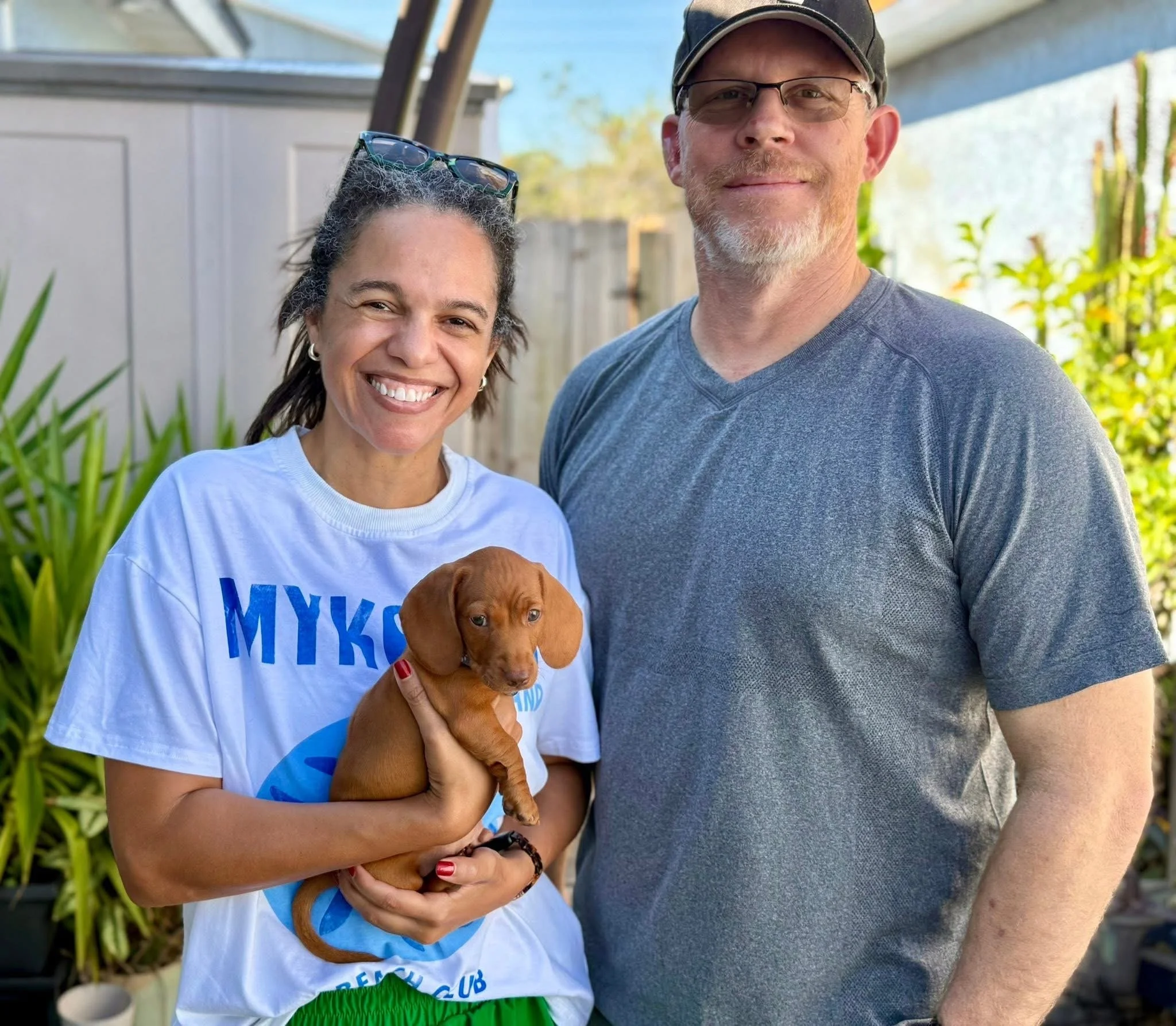 A smiling woman with short curly hair and sunglasses on her head, holding a small brown puppy, standing next to a man with a beard, glasses, and a cap, outdoors in a sunny area with greenery and a shed in the background.