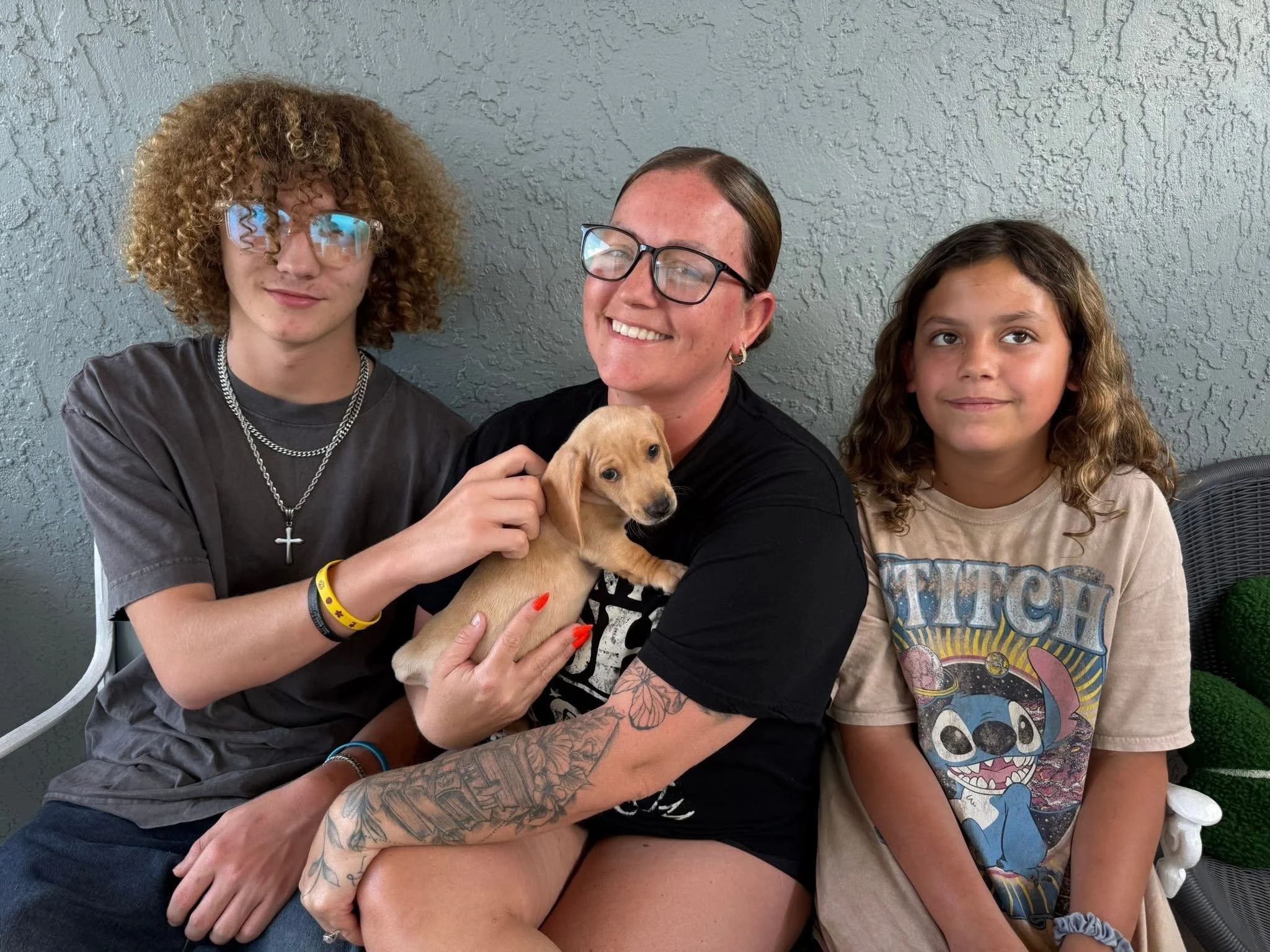 Three people and one puppy sitting together against a textured gray wall. The person on the left has curly red hair and glasses, wearing a black shirt with jewelry. The person in the middle has brown hair, glasses, earrings, and tattoos on their arms