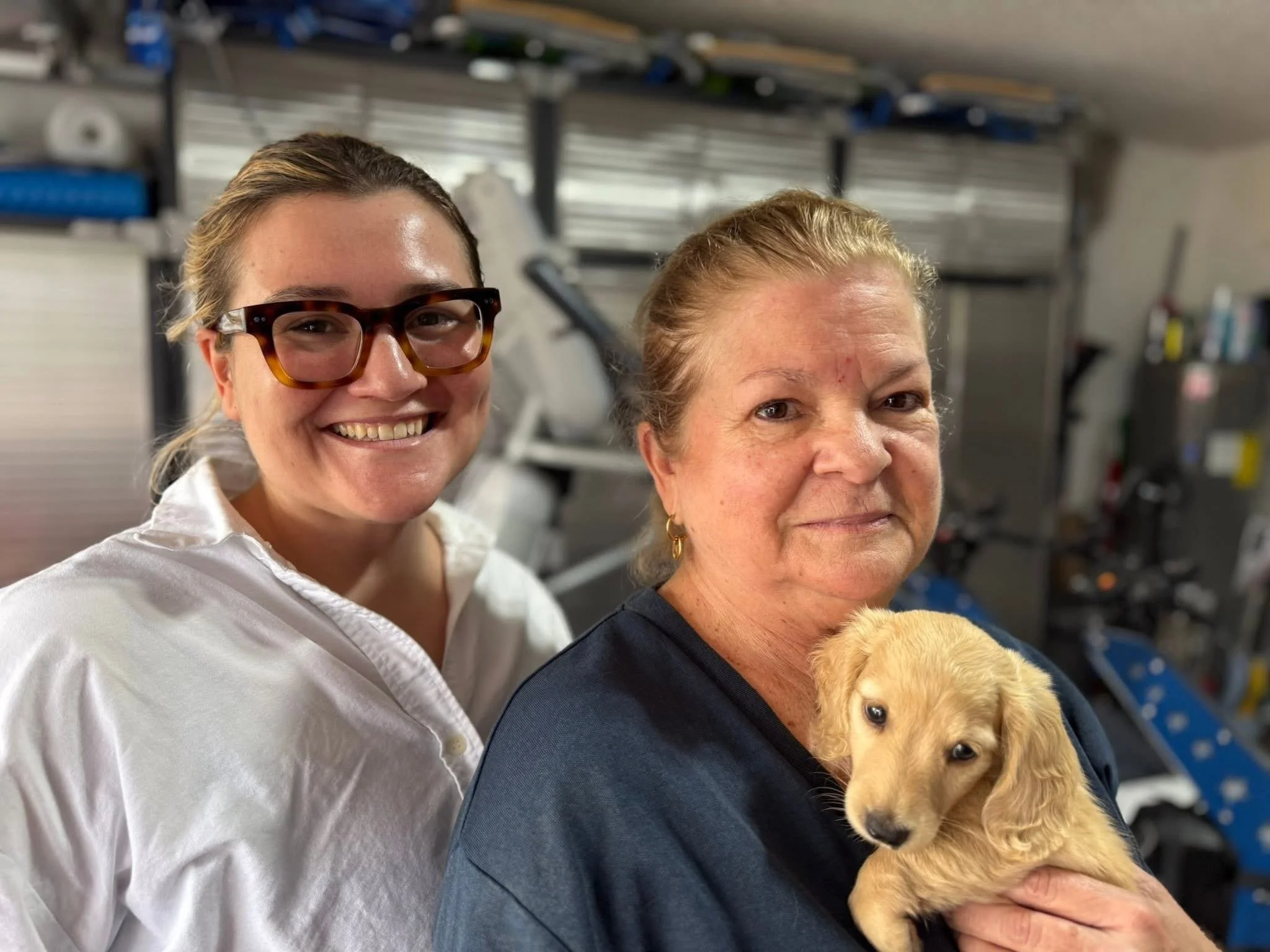 A woman with blonde hair and earrings holds a tiny yellow puppy. Another woman with glasses and a smile is beside her. They are in a room with shelves of supplies in the background.