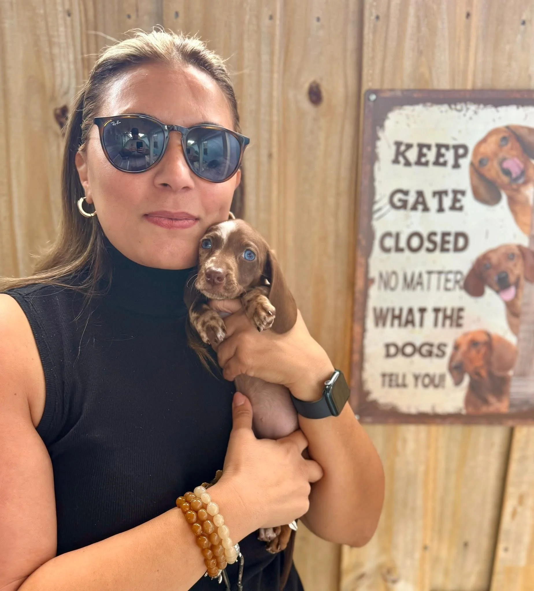 A woman with long hair wearing sunglasses, hoop earrings, and a black sleeveless top holding a small brown puppy with blue eyes in front of a wooden background with a sign that says 'Keep Gate Closed' and features images of dogs.